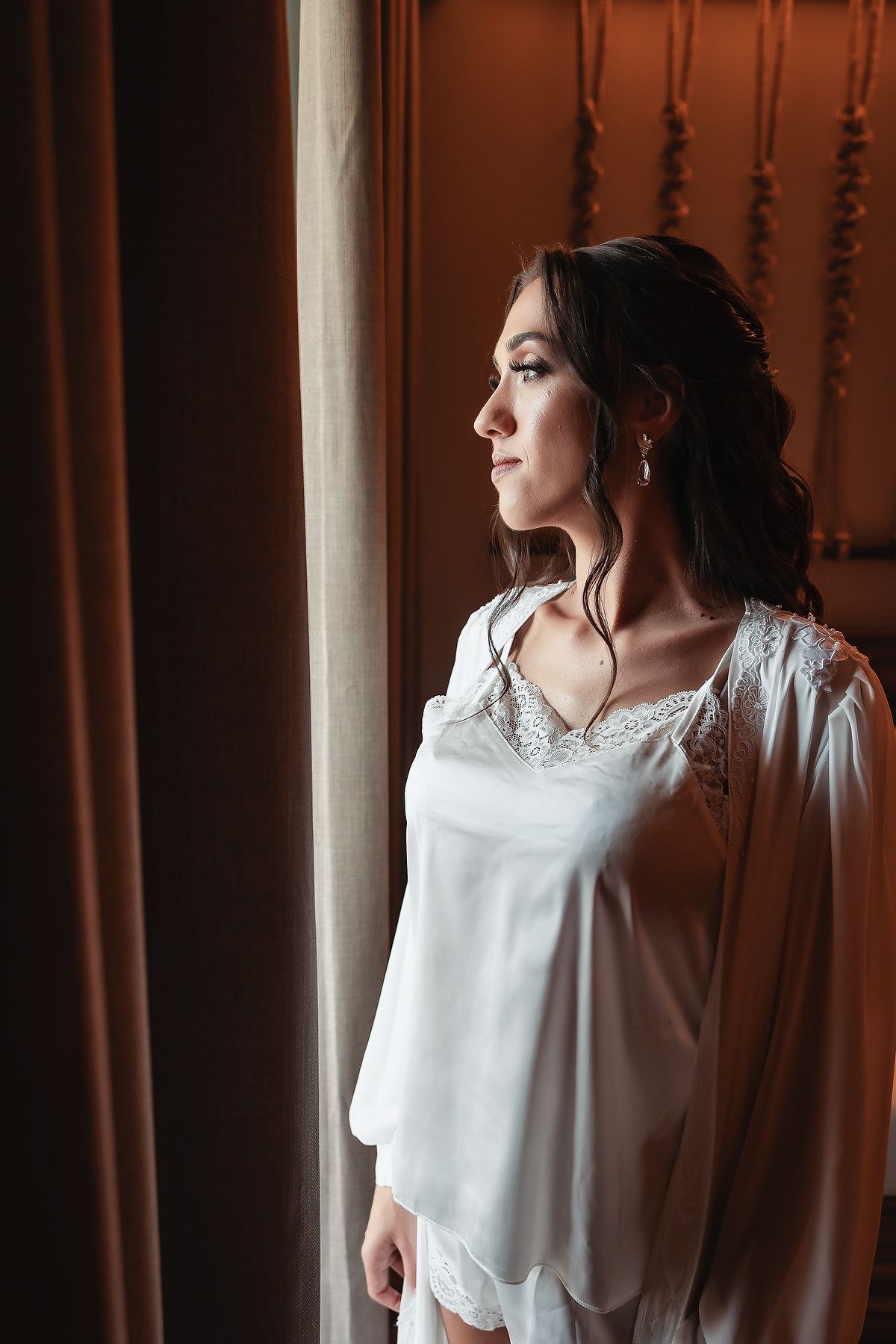 Bride in white lace robe gazing out window with elegant updo and makeup in soft warm light.