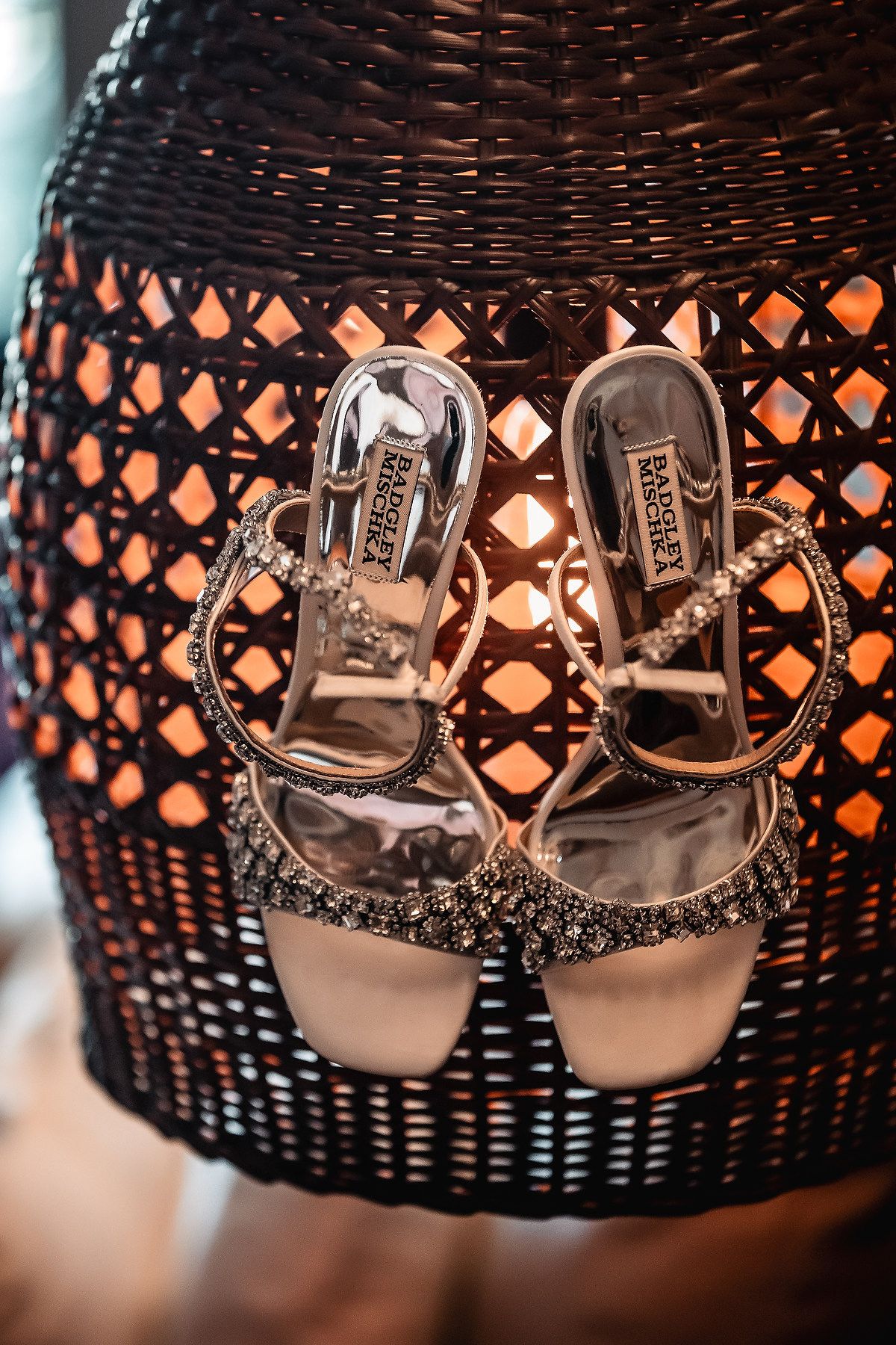 Badgley Mischka silver embellished strappy heels displayed on a dark wicker chair with warm glowing light.
