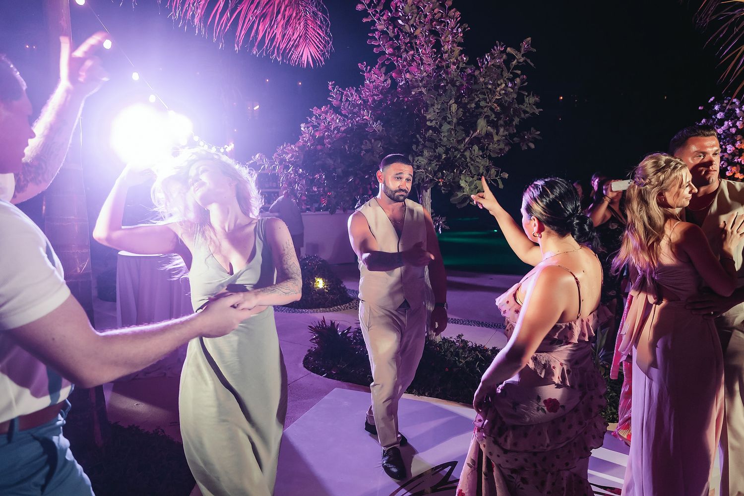 Guests dancing and celebrating outdoors at a nighttime party with bright stage lighting and palm trees.