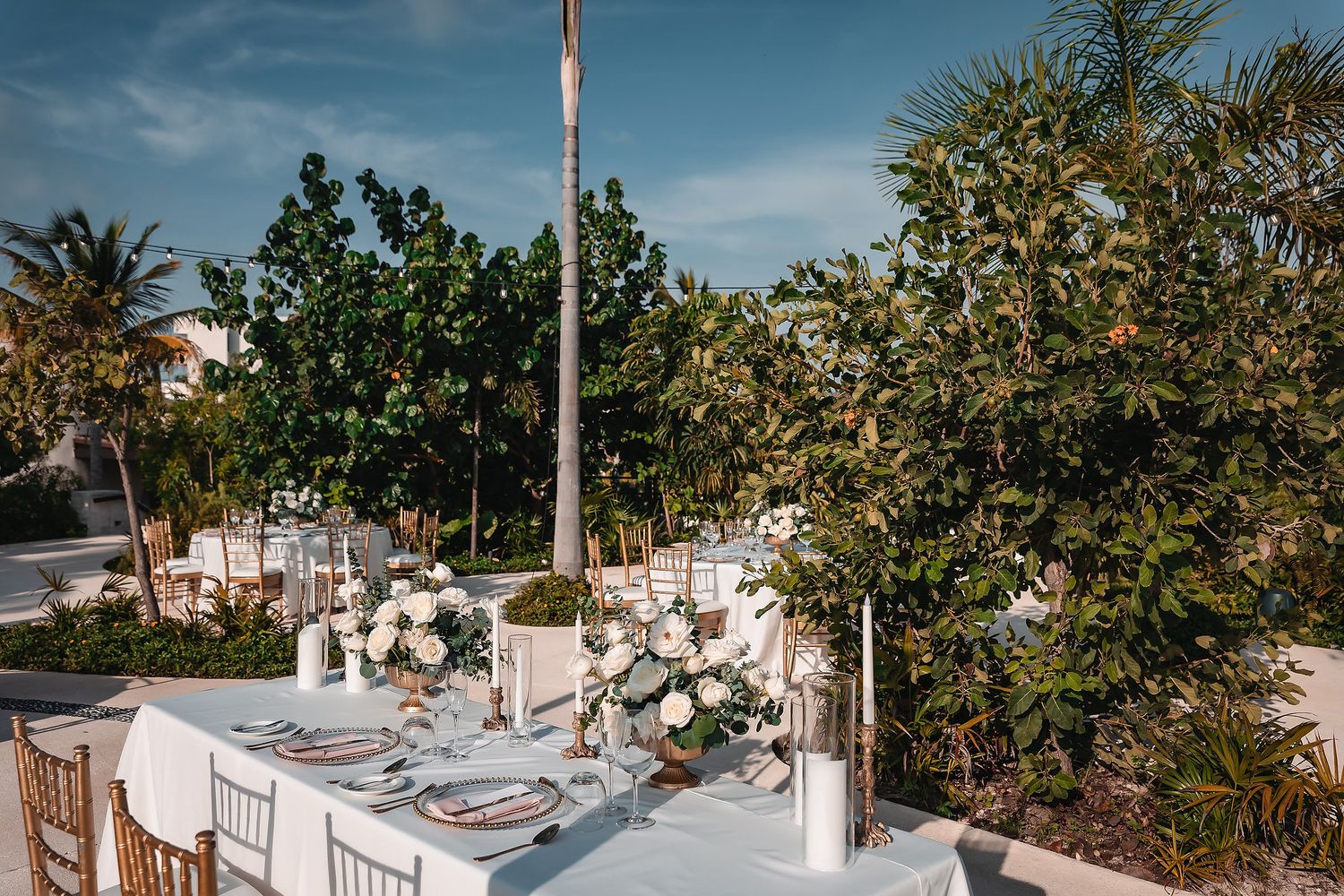 Elegant outdoor wedding reception table with white floral centerpieces, gold chairs, and lush tropical greenery under blue skies.