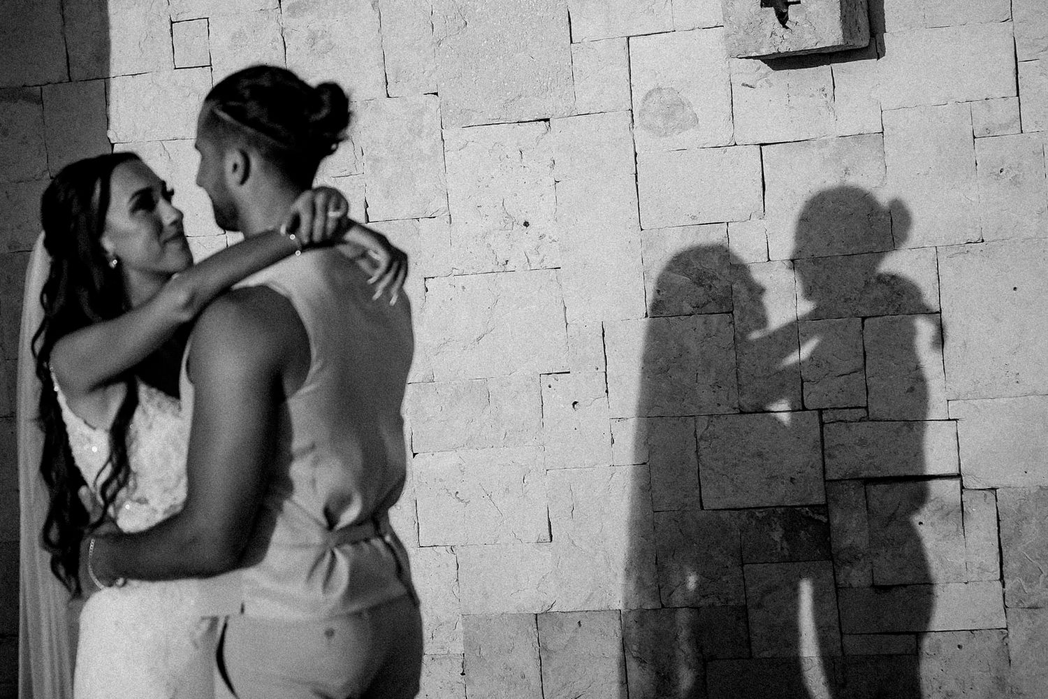Black and white wedding photo of couple embracing against stone wall with romantic shadows cast behind them.