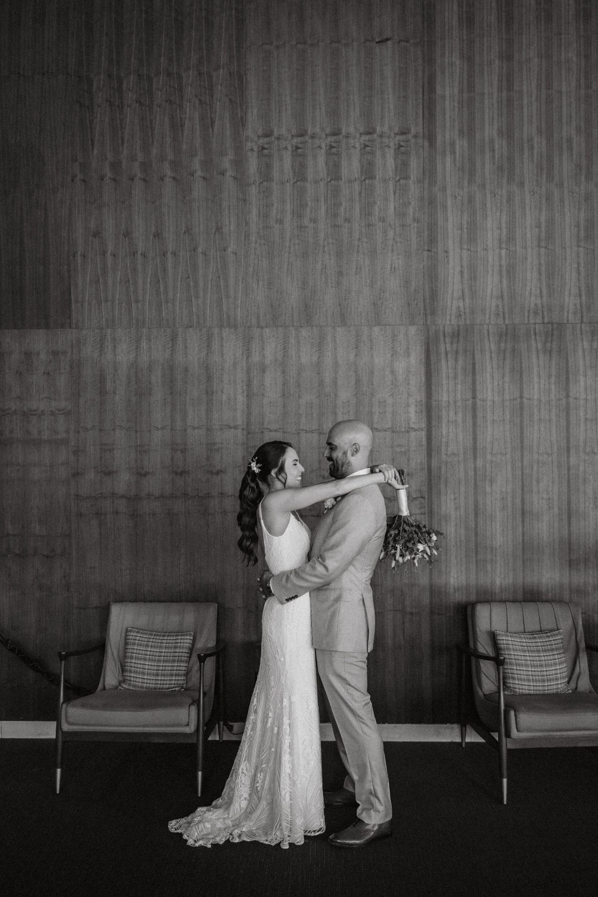 Black and white photo of bride and groom embracing in front of wood panel wall with chairs beside them.