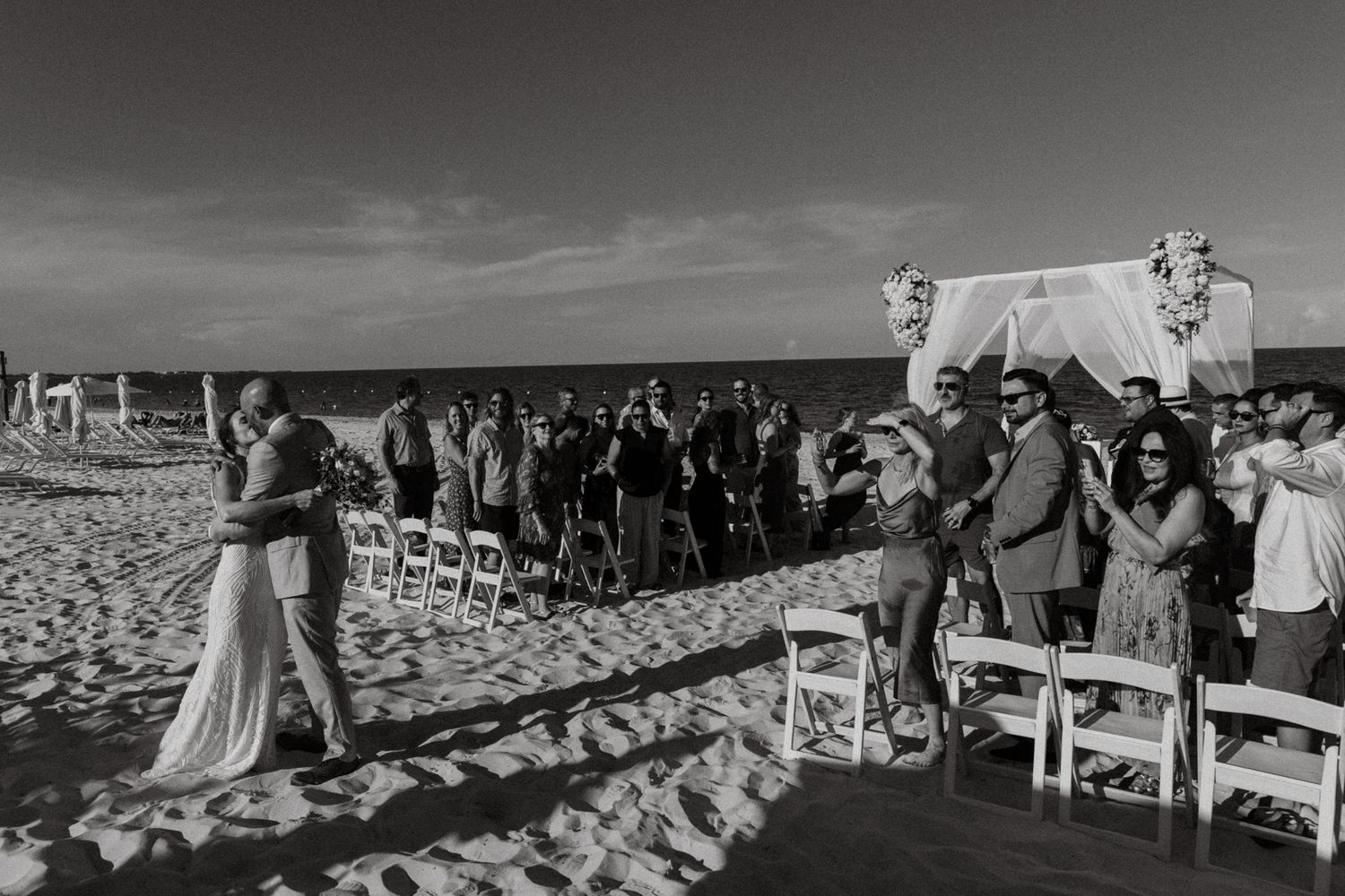 Newlyweds share first kiss on beach as wedding guests celebrate around them at outdoor ceremony.