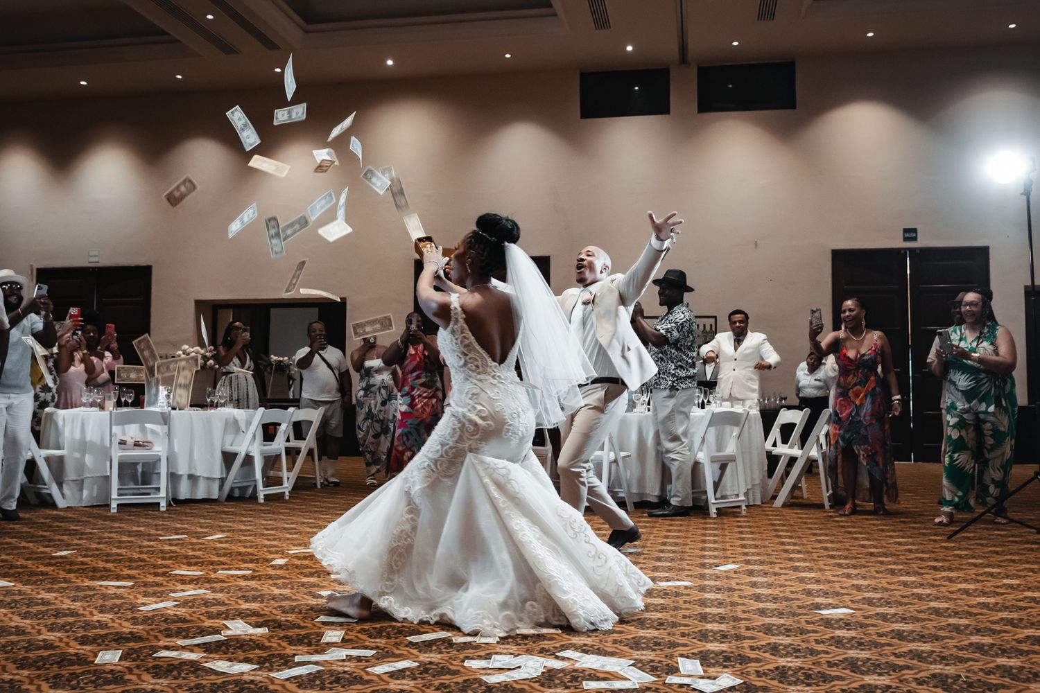 Bride in a white lace gown celebrates on the dance floor as money flies through the air at her reception.
