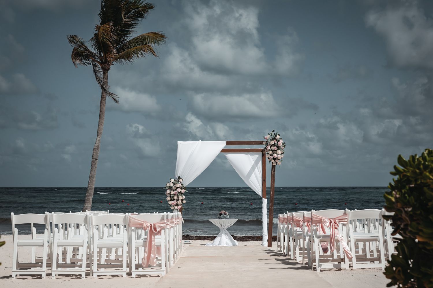 Beach wedding setup with white chairs, floral arch, and palm tree overlooking the ocean under a cloudy sky.