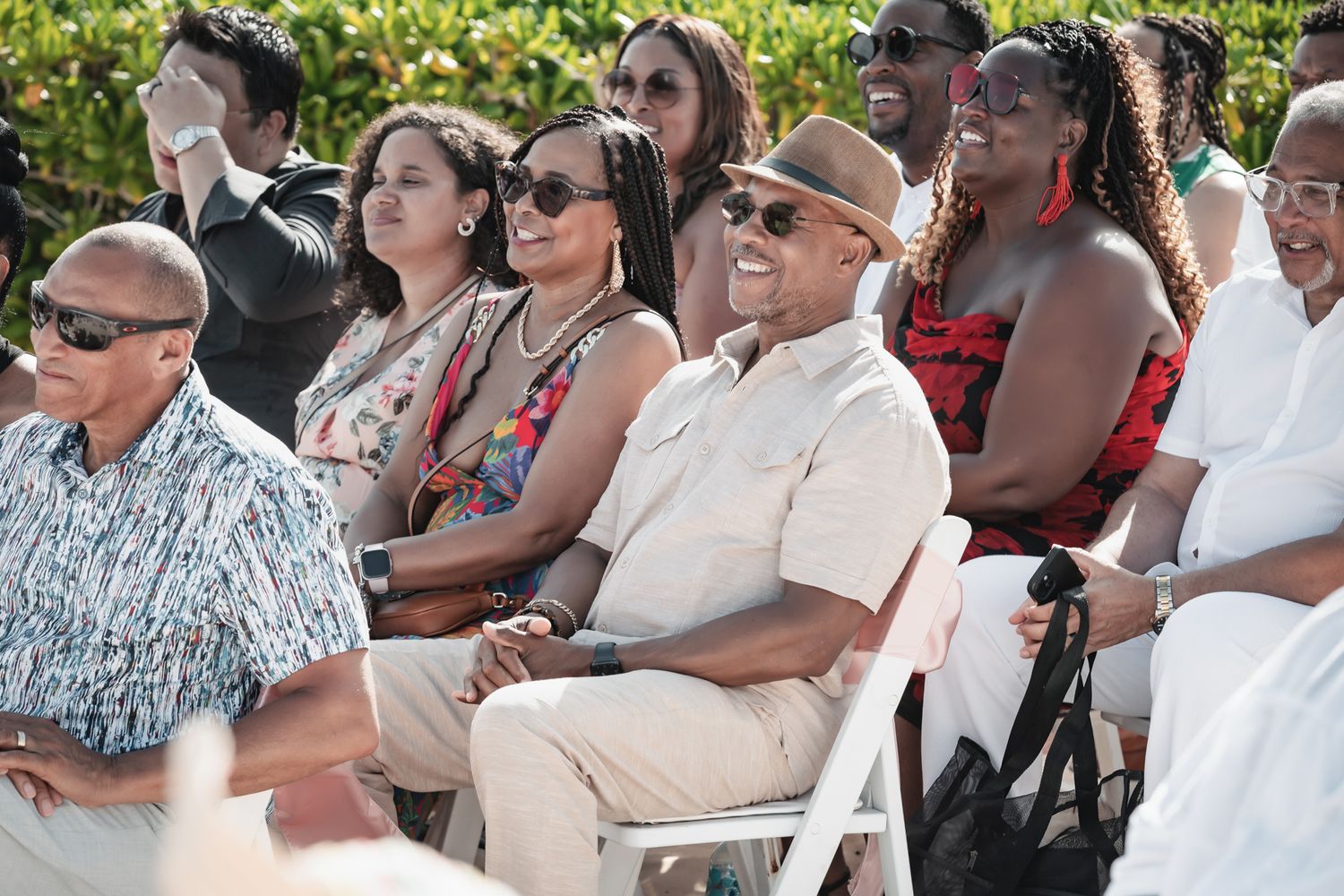Diverse group of smiling guests seated outdoors at a sunny celebration event, dressed in colorful attire.