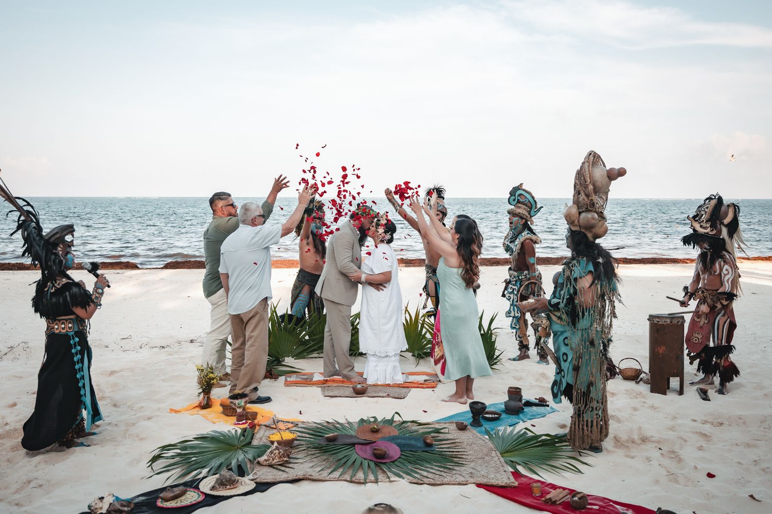 Couples beach wedding ceremony with Mayan warriors in traditional costumes tossing red rose petals at sunset.