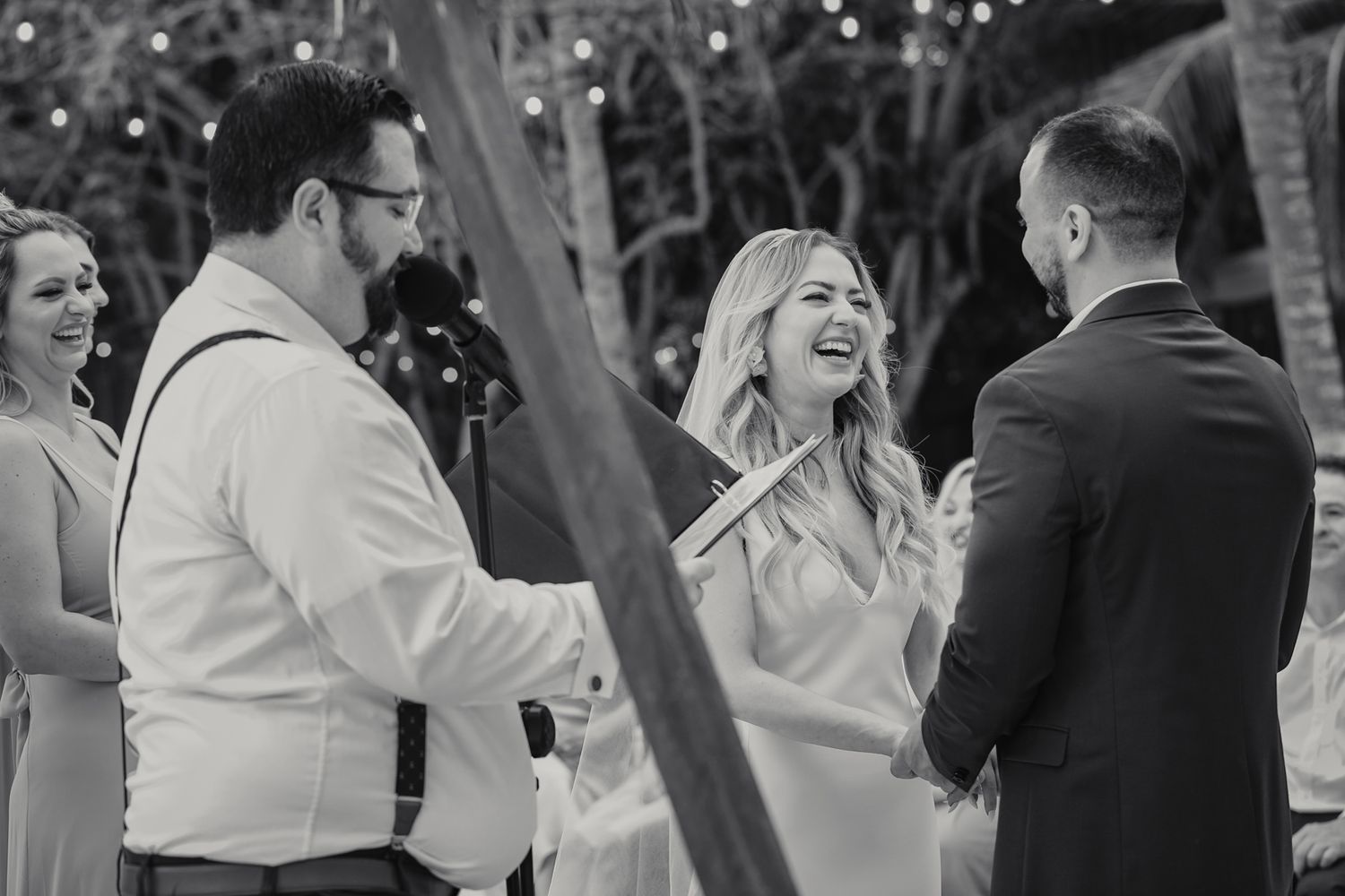 Bride laughs joyfully during outdoor wedding ceremony while exchanging vows with officiant and groom in black and white photo.