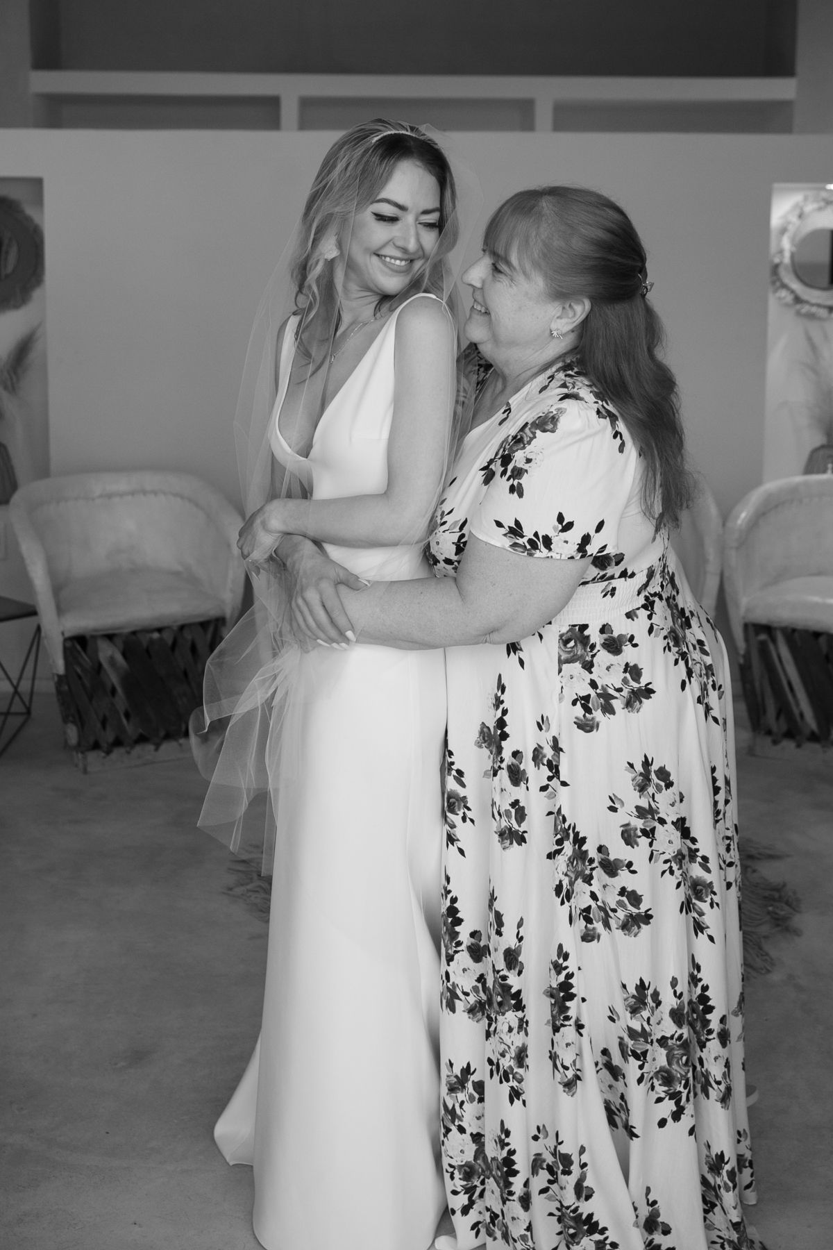 Bride in white gown embracing older woman in floral dress during wedding preparations in black and white.