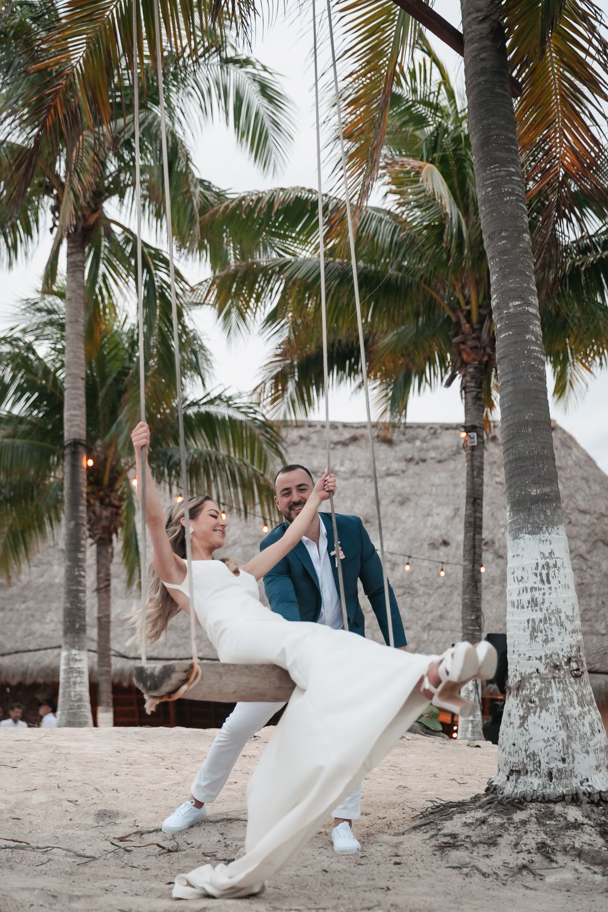 Bride on a beach swing with groom pushing her among tropical palm trees at sunset wedding.