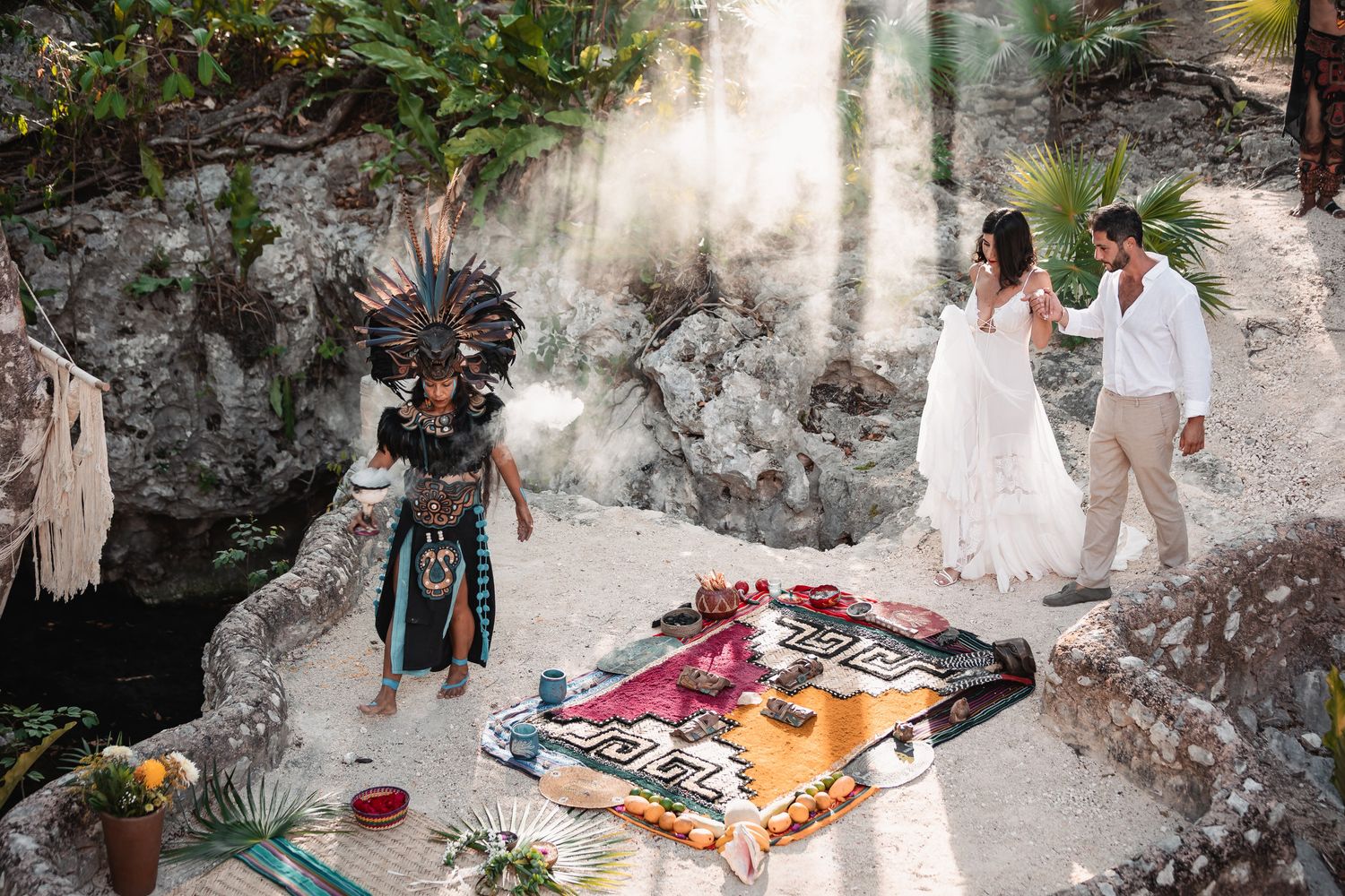 Mayan ceremony at a cenote with a costumed officiant blessing a couple in wedding attire on a colorful textile.