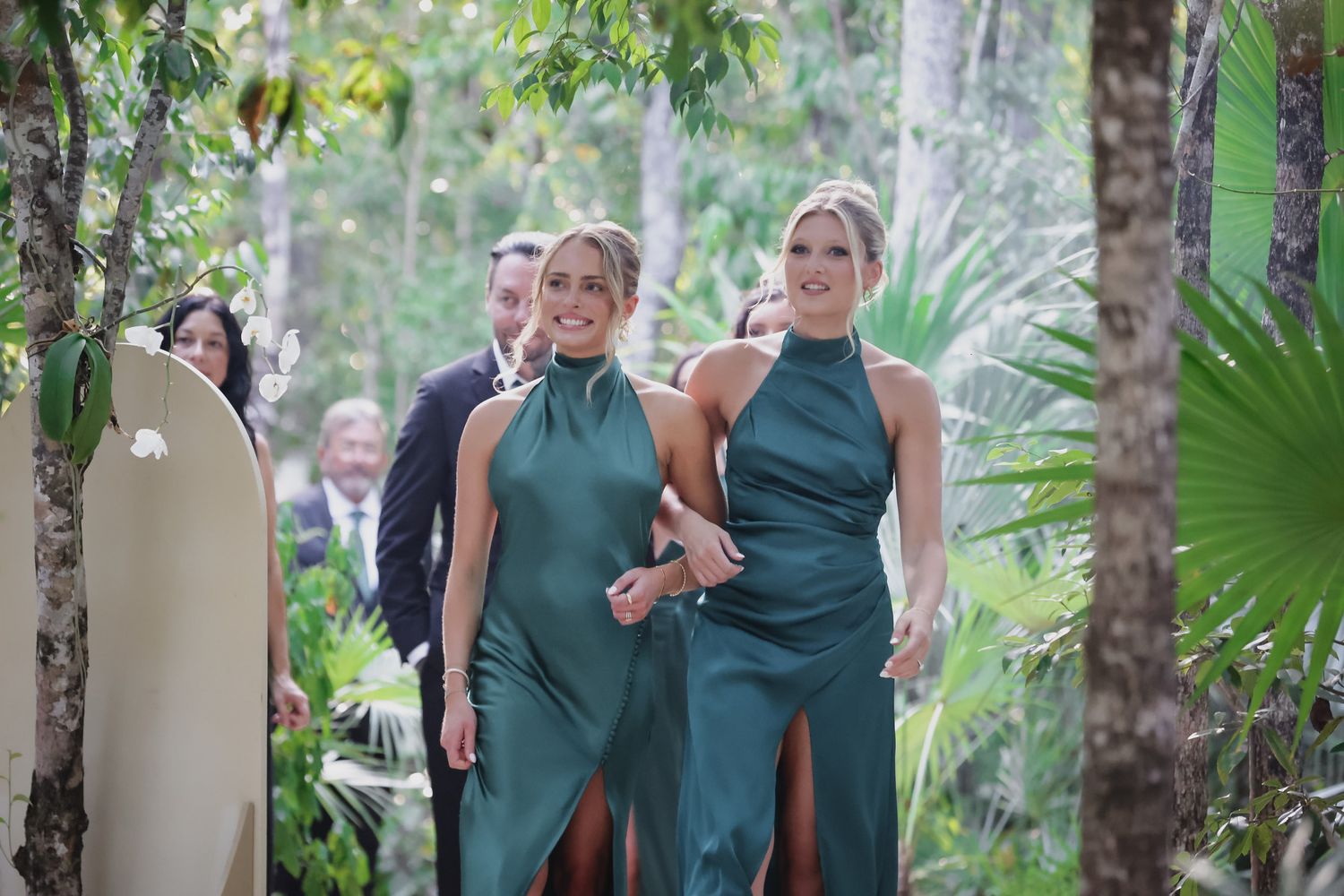 Two bridesmaids in elegant green halter dresses smile walking through tropical jungle wedding.