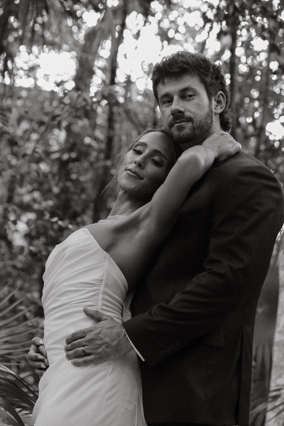 Black and white wedding photo of bride and groom embracing in lush forest setting, bride in white gown resting head on groom.
