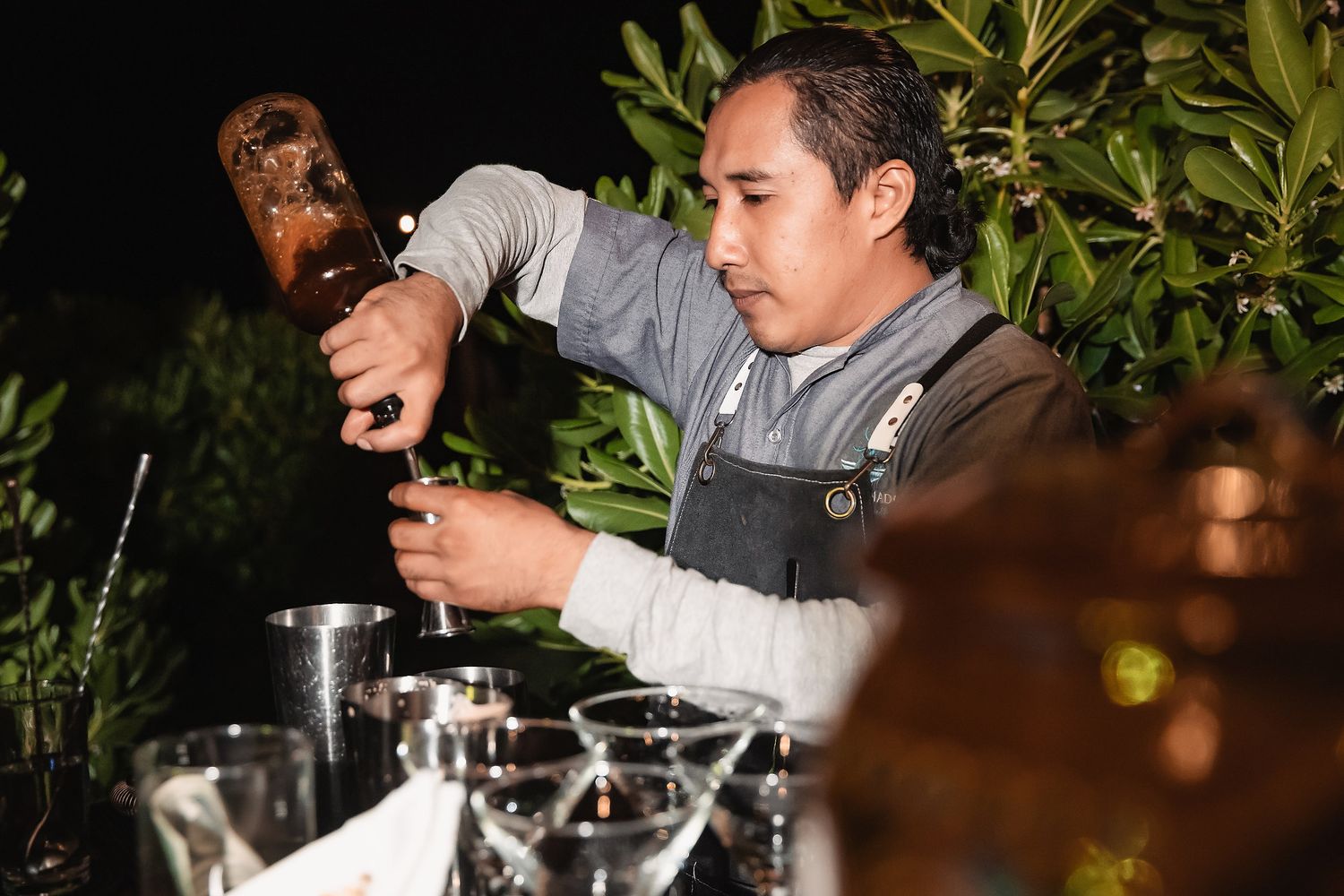 A bartender pours a drink at an outdoor night event, surrounded by glassware and bar tools on a table.