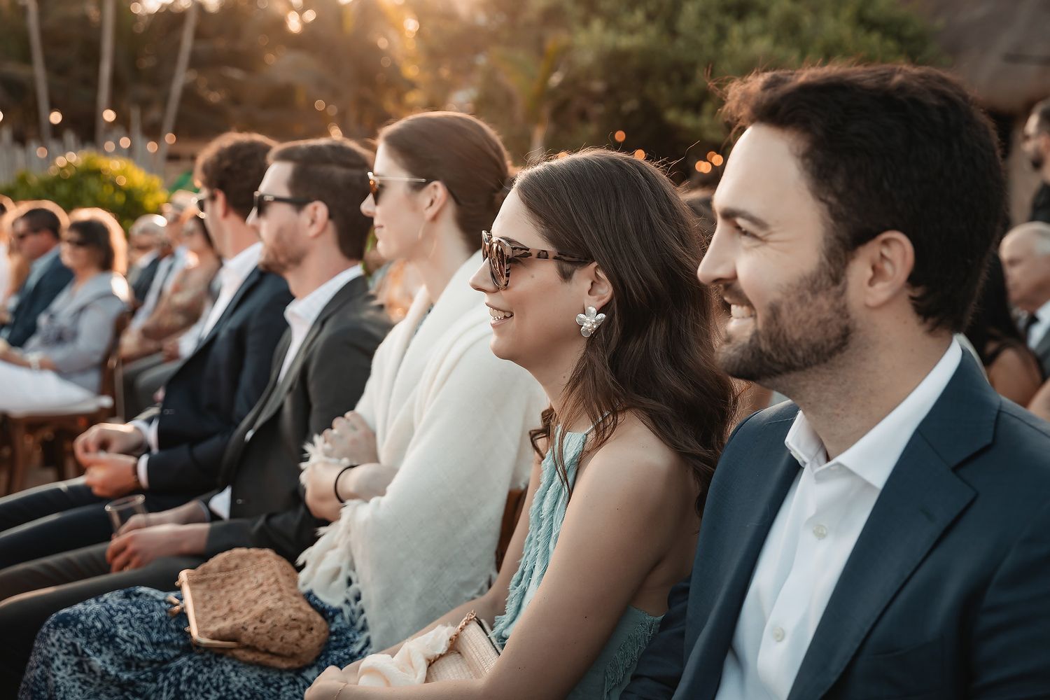 Guests smiling and seated outdoors at a wedding ceremony during golden hour sunset.