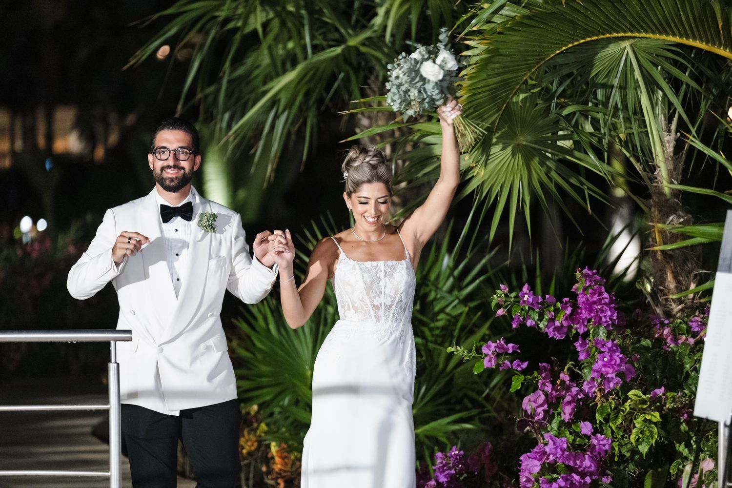 Bride and groom celebrate at outdoor wedding ceremony, bride raising bouquet, surrounded by tropical flowers.