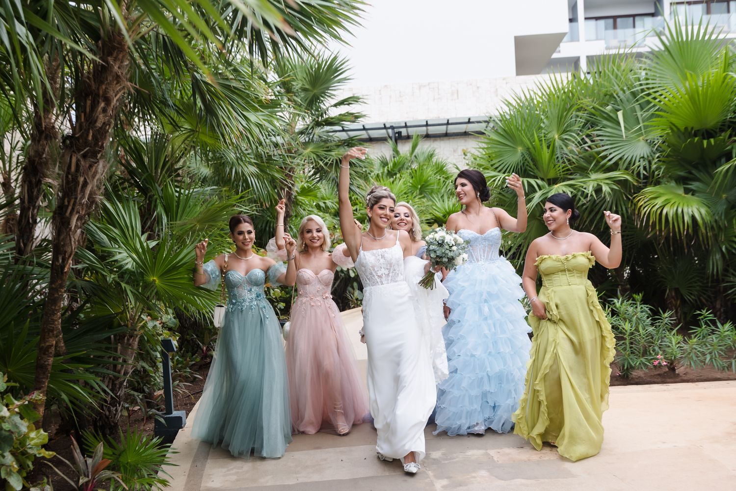Bridesmaids in colorful pastel gowns celebrate with bride outdoors among tropical palm trees.