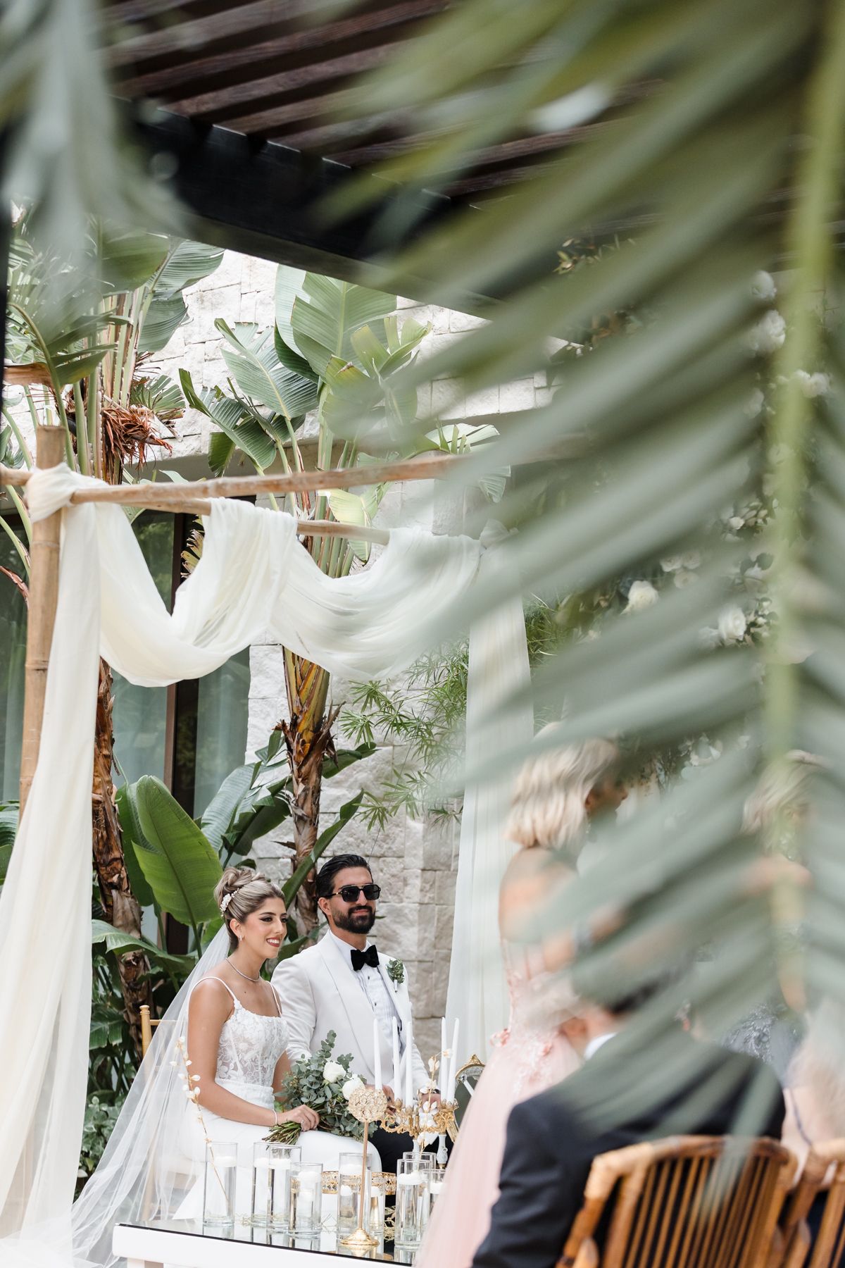 Bride and groom stand under draped white chuppah surrounded by lush tropical greenery during outdoor wedding ceremony.
