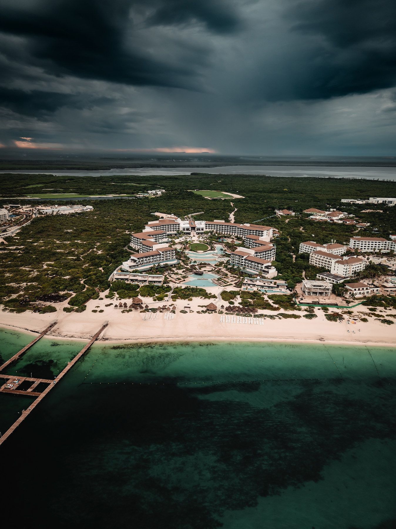 Aerial view of a luxury beach resort surrounded by lush greenery and turquoise waters under dramatic storm clouds.