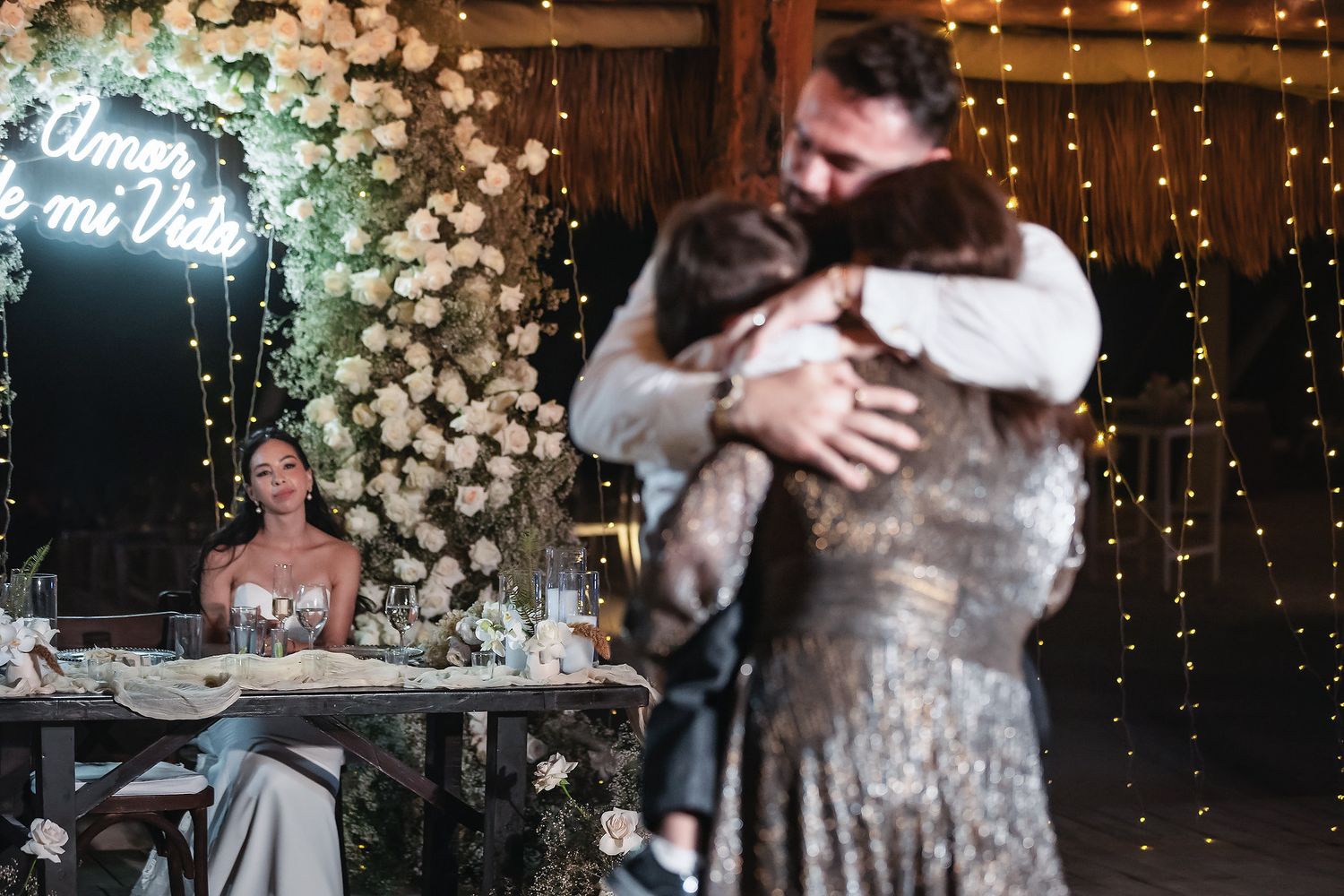A couple shares an emotional hug at a romantic wedding reception with floral decor and neon sign in background.