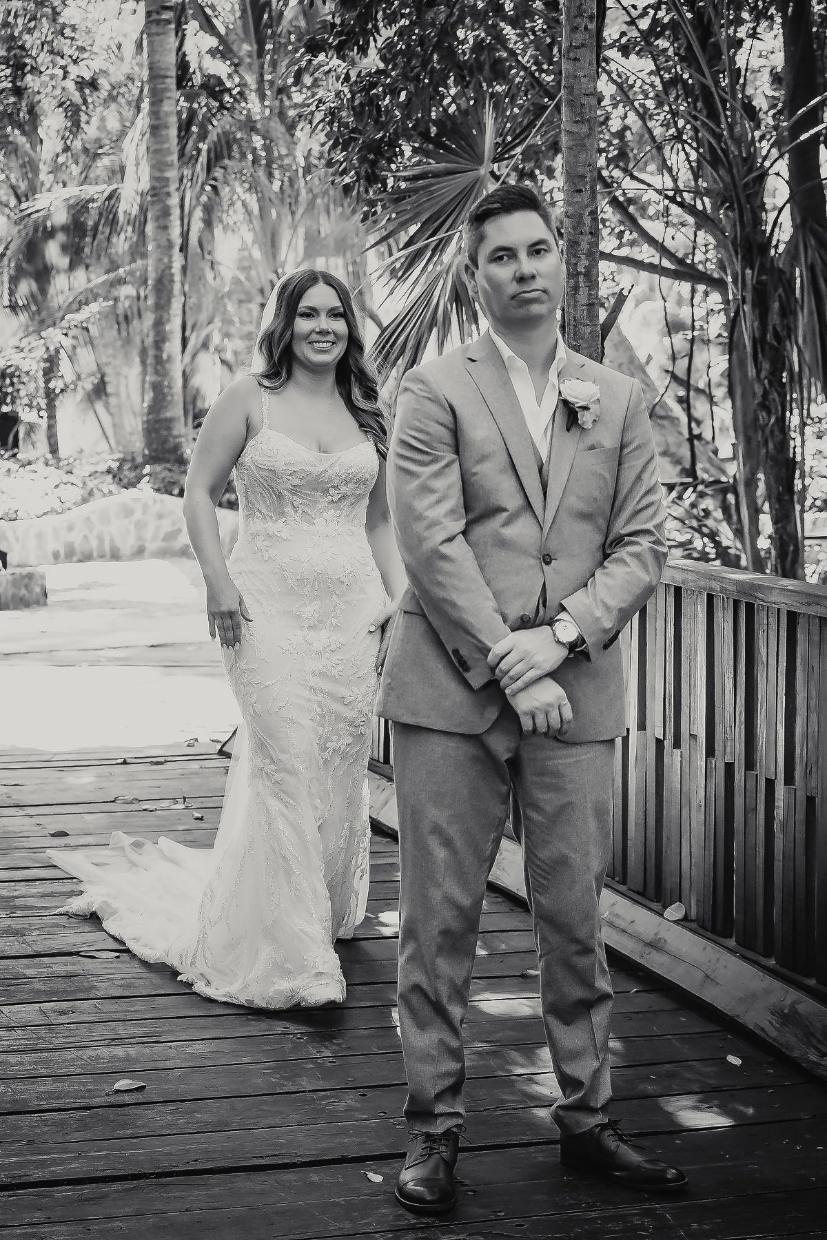 Black and white wedding photo of bride in lace gown and groom in tan suit on wooden deck surrounded by tropical trees.