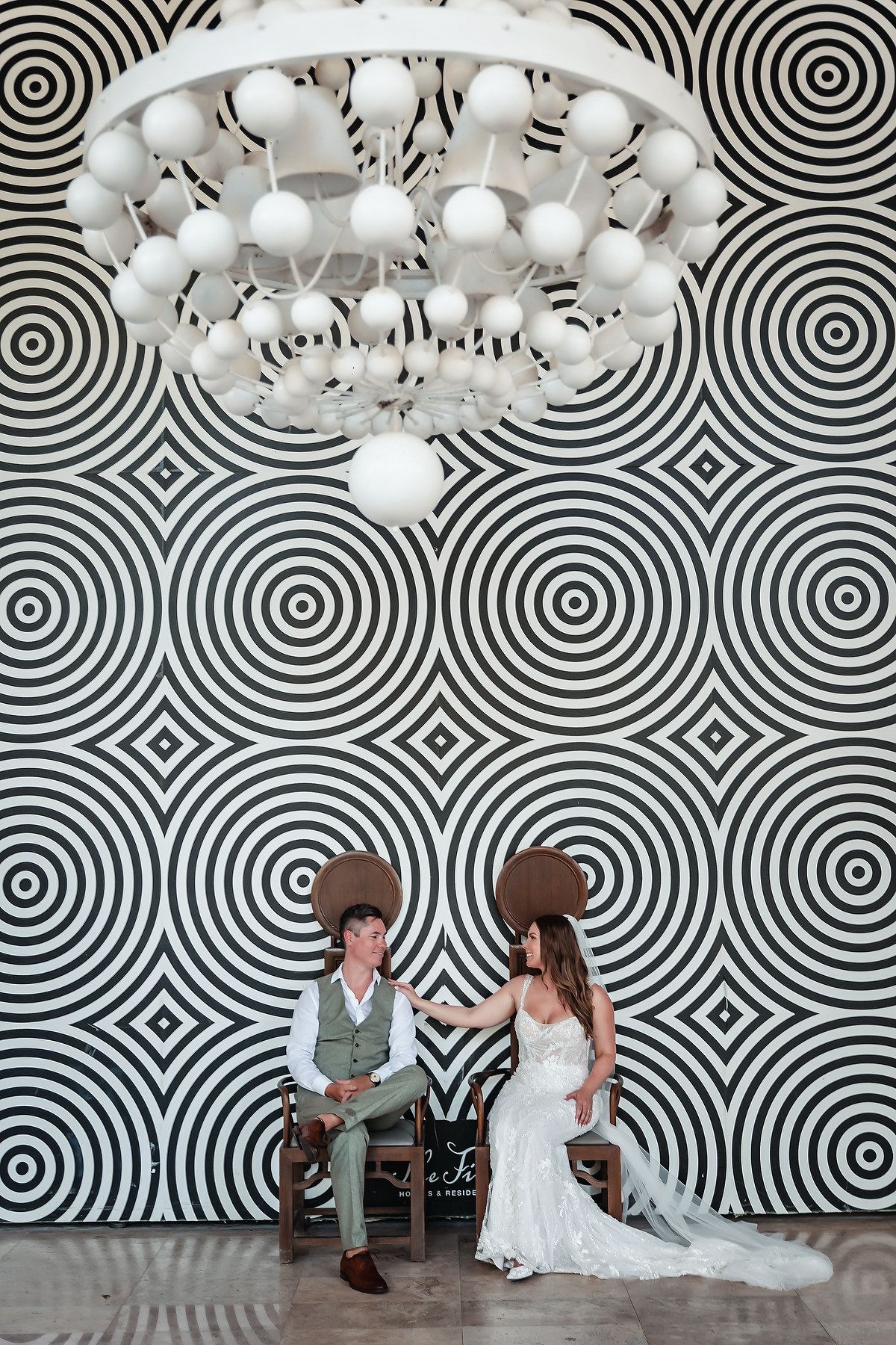 Bride and groom in wide-brim hats sit on chairs before a bold black-and-white circular patterned wall with chandelier.