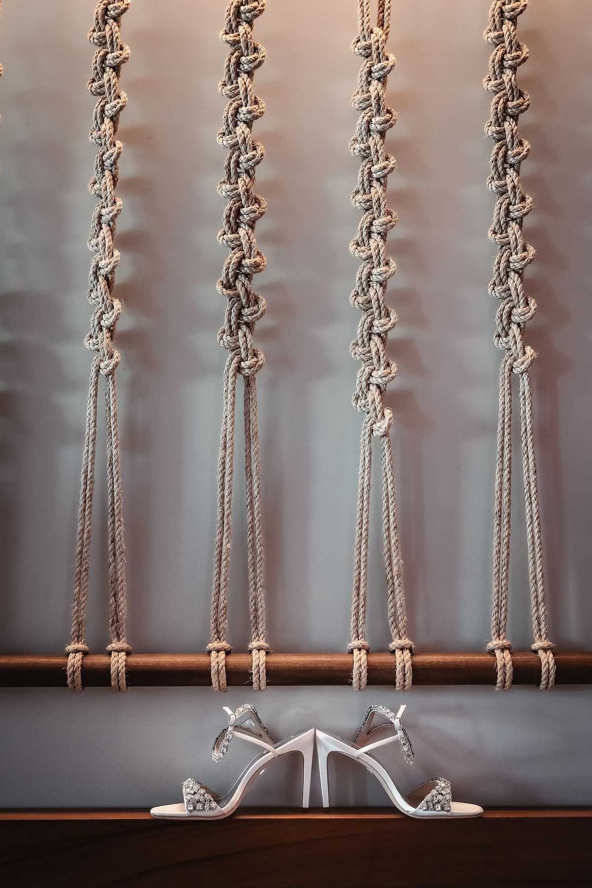 Elegant white bridal heels displayed on a wooden shelf against a decorative macrame rope backdrop.