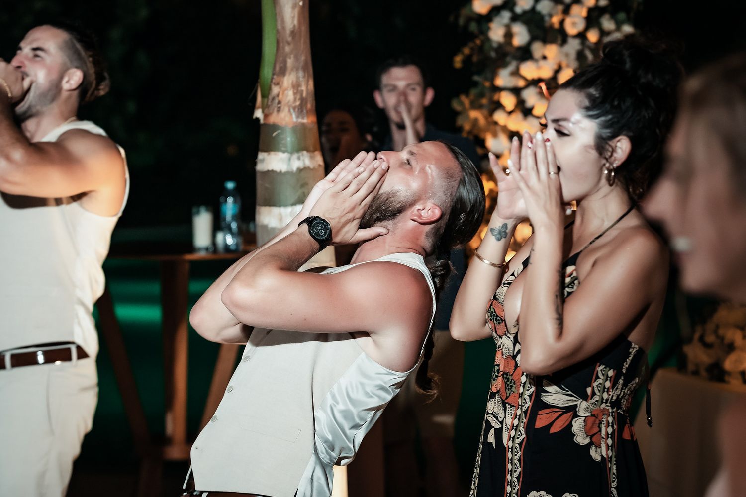Two guests at an outdoor evening party react with surprise, hands raised to their faces, at a tropical venue.