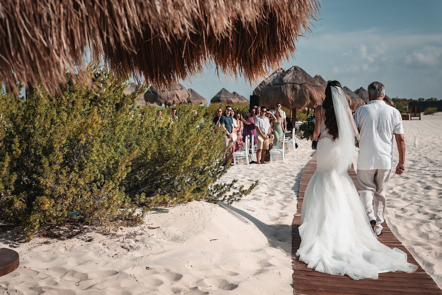 Bride and groom walking on a wooden boardwalk toward a beach wedding ceremony under a thatched palapa.