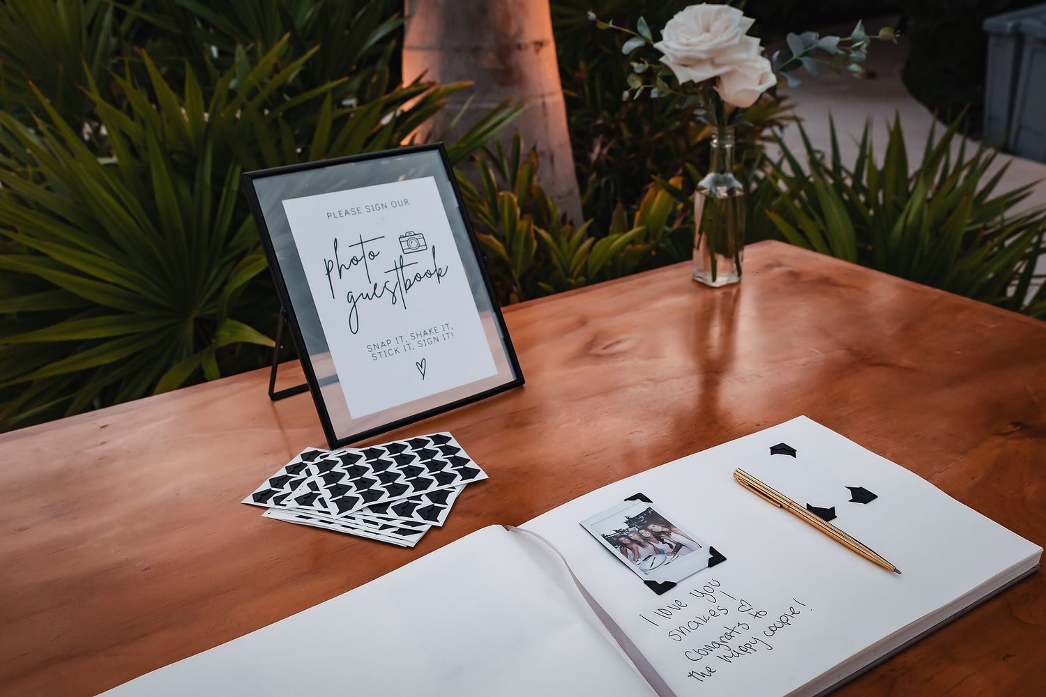 Wedding guest book table with framed photo guestbook sign, polaroid photos, and open journal on wooden surface.