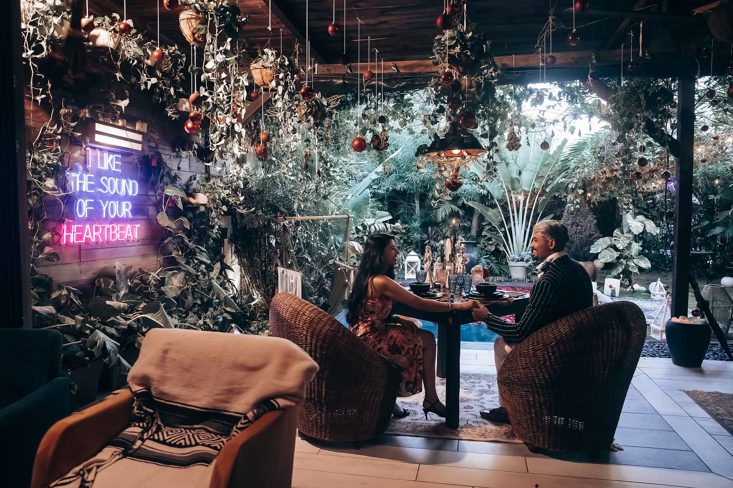 Couple dining in a lush indoor garden restaurant with neon sign reading love quote and hanging floral decorations.