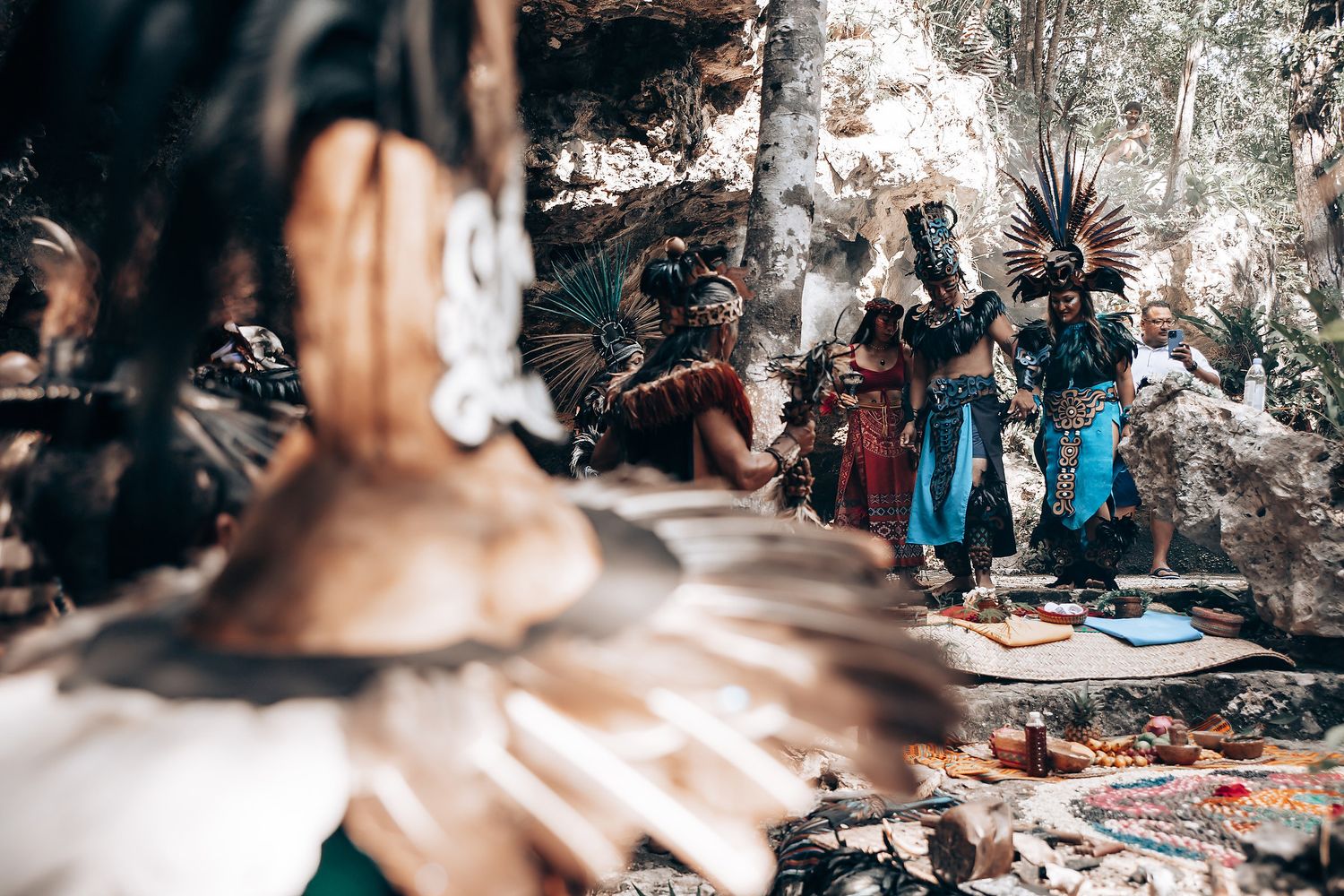 Group of performers in traditional Aztec ceremonial attire with feathered headdresses gathered in a rocky jungle setting.