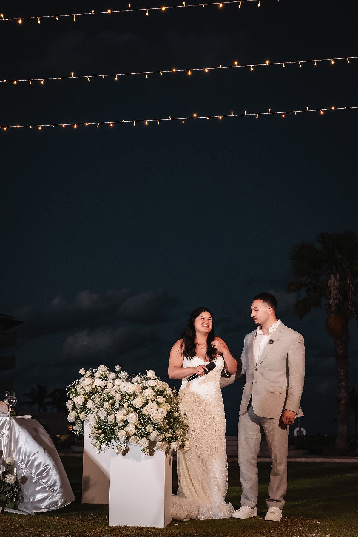 Bride and groom stand together at night wedding reception with white floral arrangement and string lights overhead.