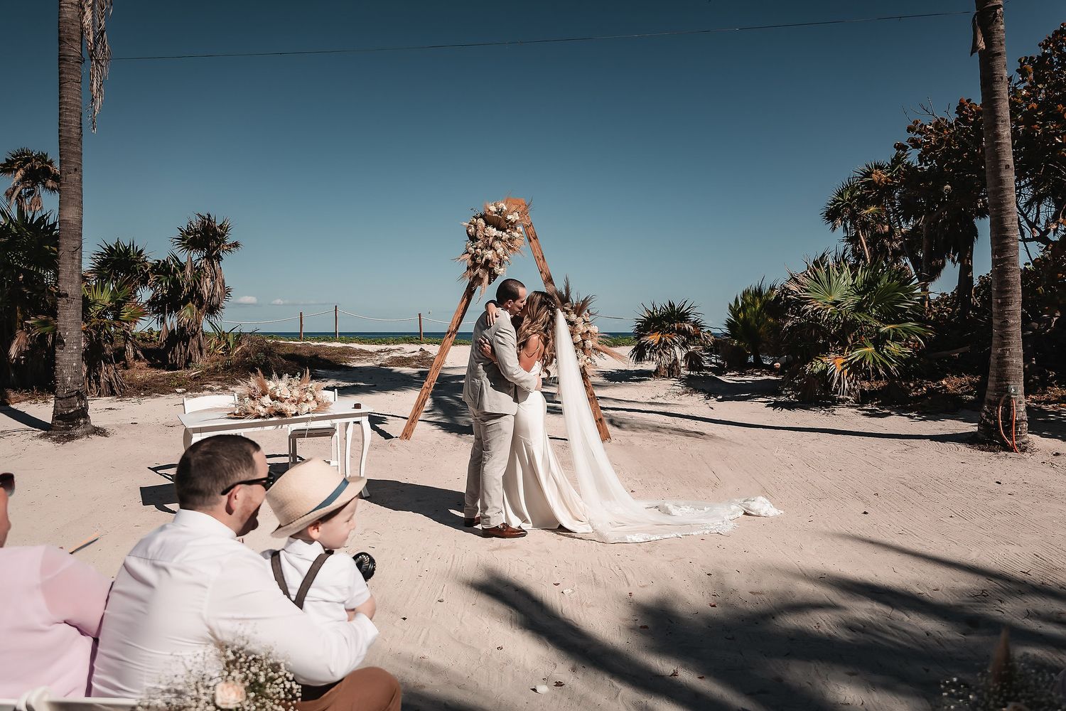 Couple exchanging vows at a beach wedding ceremony surrounded by palm trees and sandy shores on a sunny day.