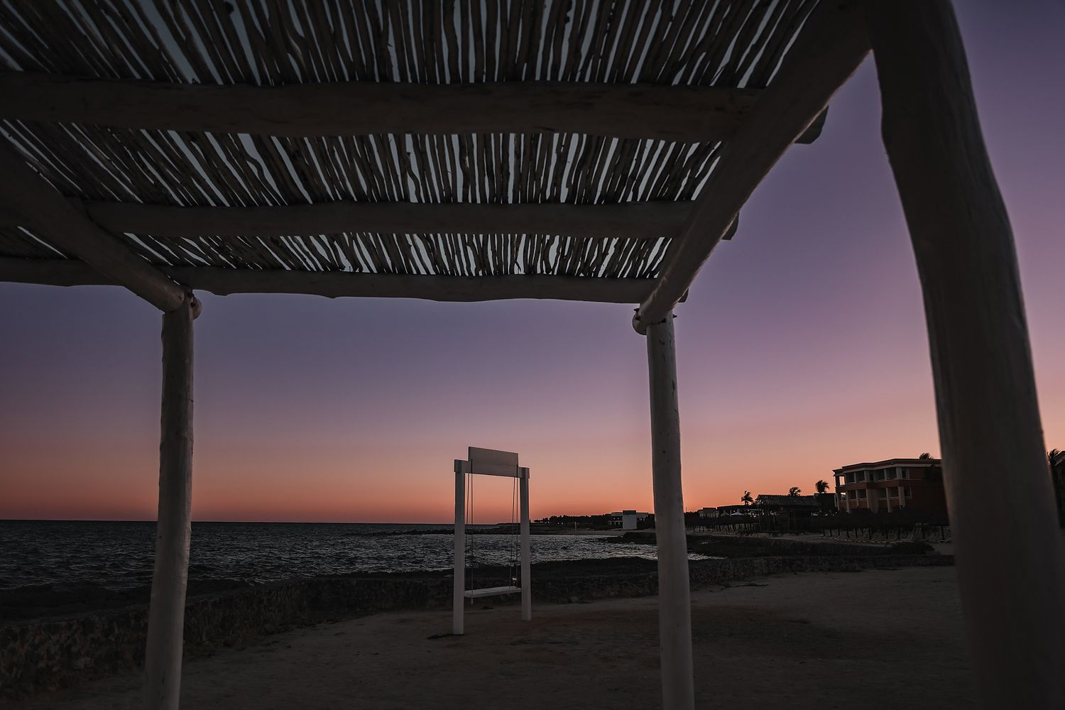 Beachside pergola silhouette at twilight with purple and pink sunset sky over ocean waters.