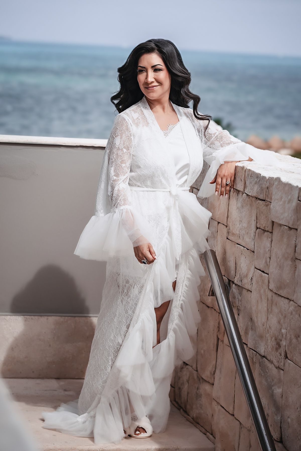Bride in white lace robe posing on a terrace with ocean views.