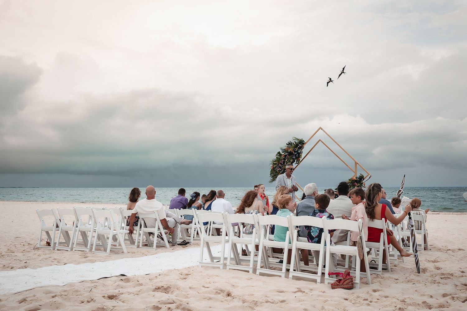 Beach wedding ceremony with guests seated in white chairs facing a geometric arch on white sand under cloudy skies.
