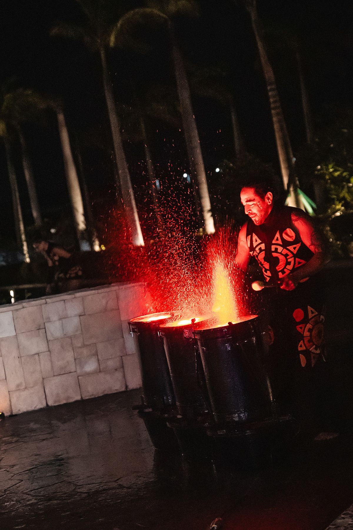 A performer drums intensely as red sparks fly from drums during a dramatic nighttime fire show outdoors.
