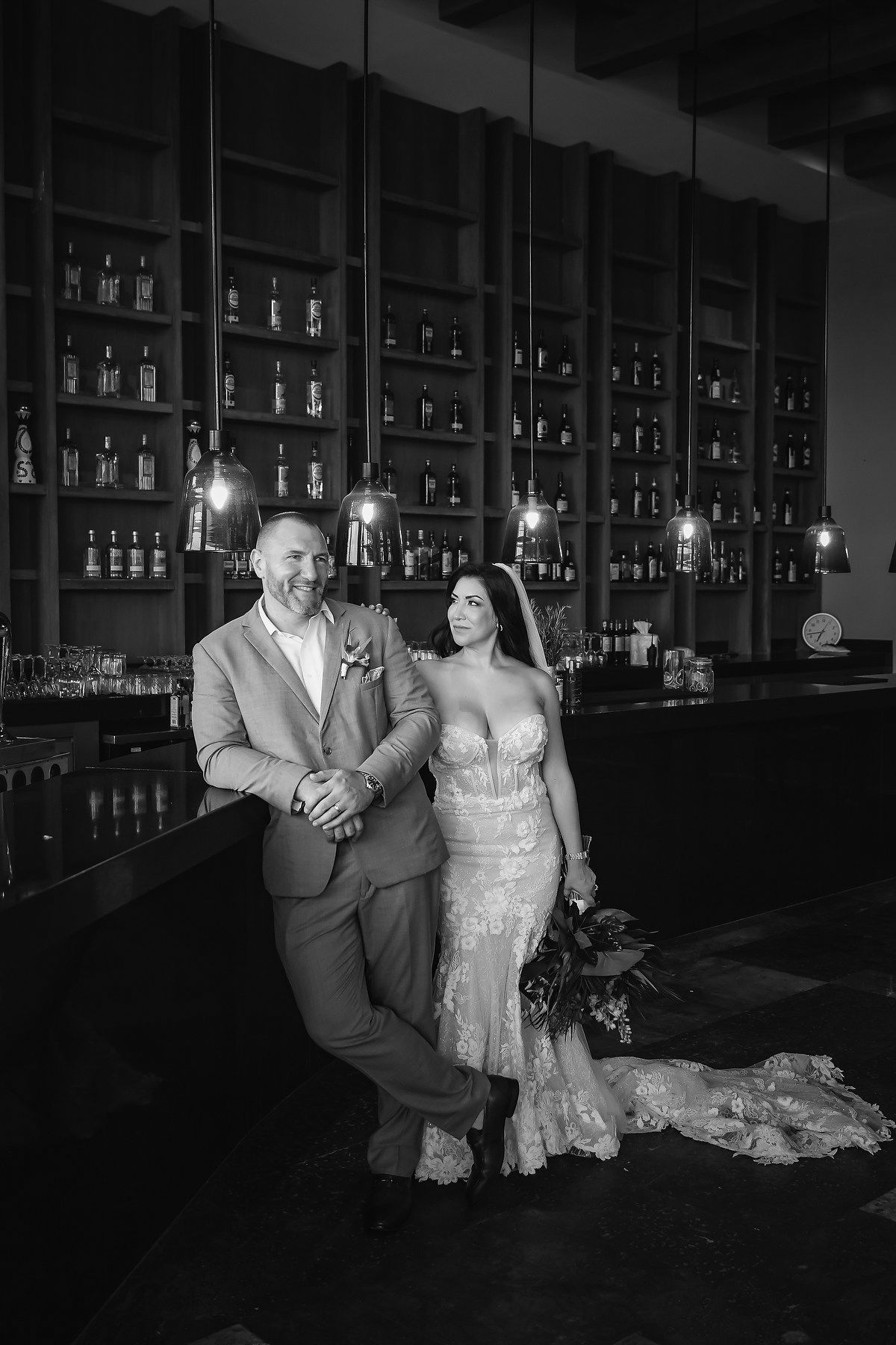 Bride and groom pose elegantly at a bar in black and white wedding portrait with bottle-lined shelves behind them.