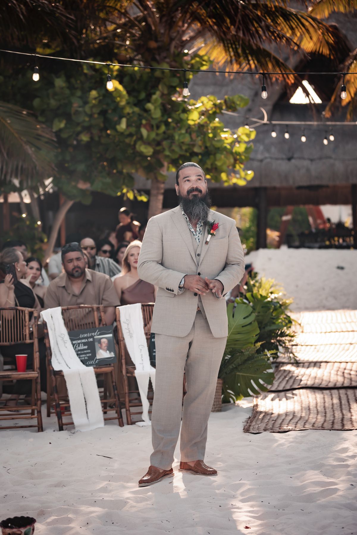 Bearded groom in tan suit with red boutonniere stands at outdoor beach wedding ceremony with string lights and tropical palms.