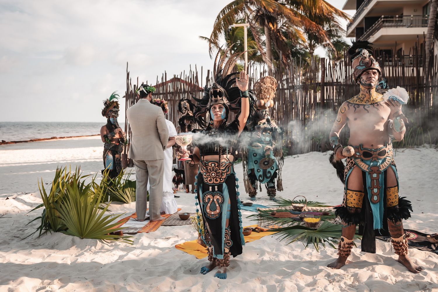 Mayan ceremony performers in traditional costumes on a beach with smoke and tropical decor.