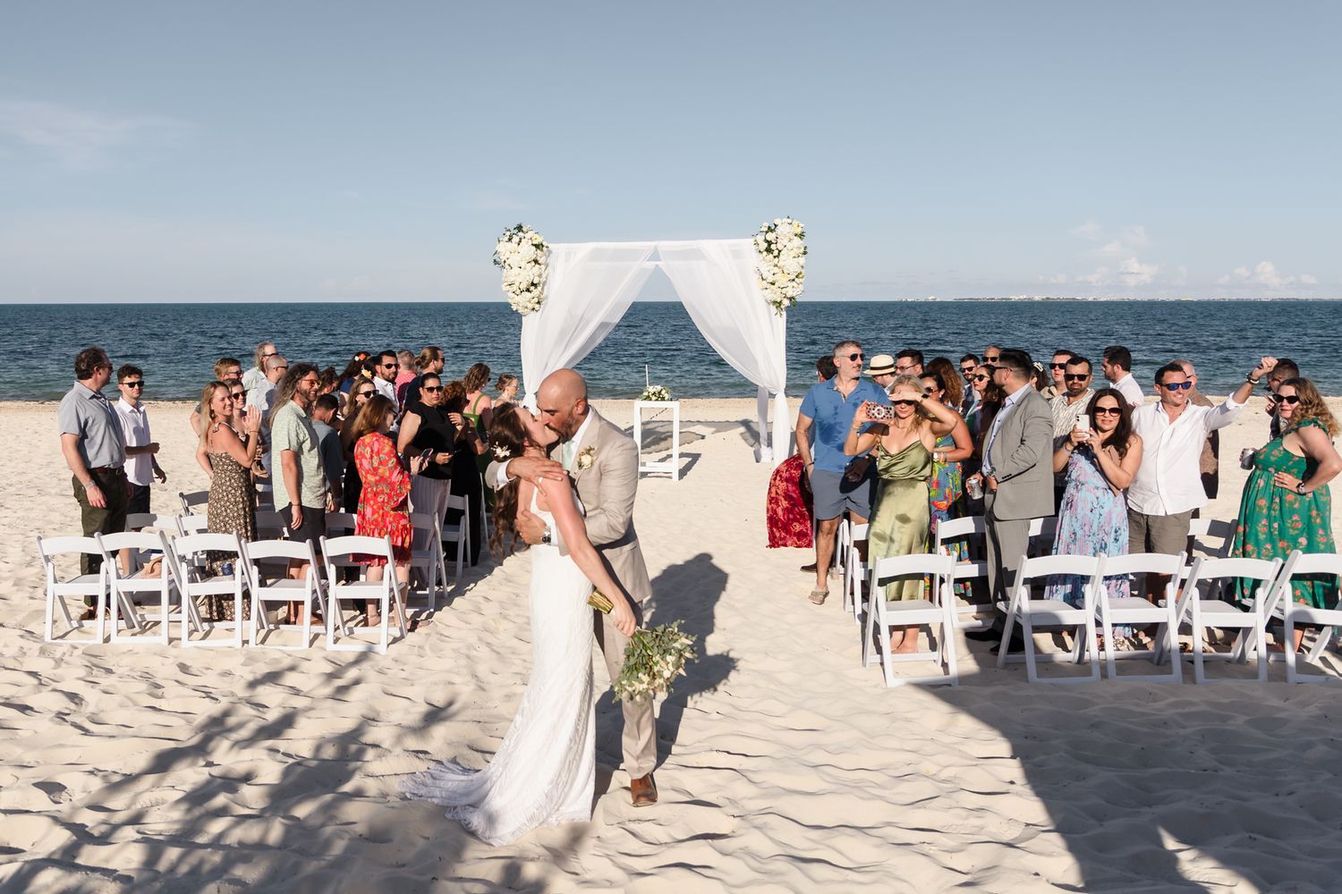 Bride and groom share a kiss at a romantic beach wedding ceremony with white chairs and floral arch.