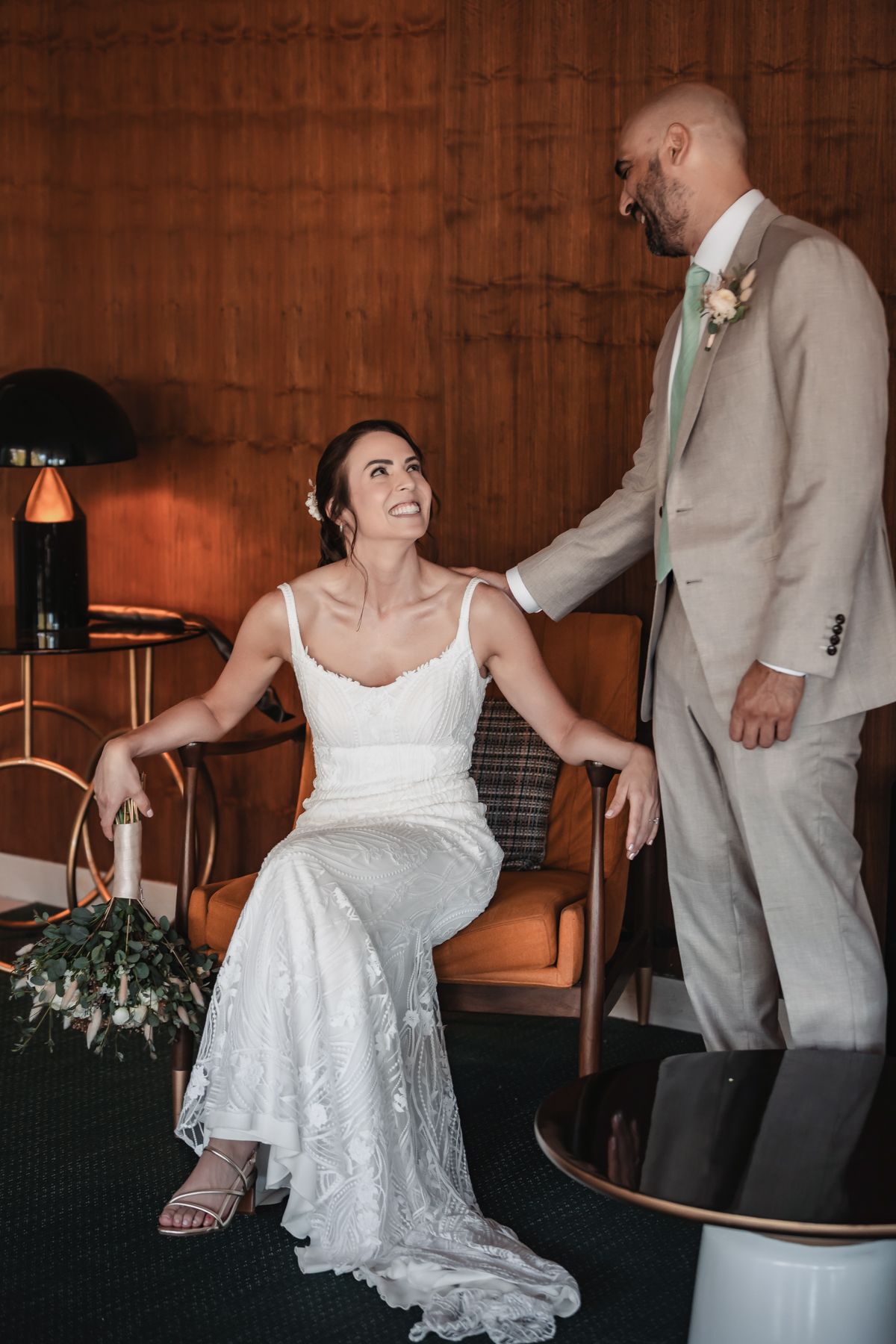 Bride in white lace gown sits smiling at groom in tan suit inside a warm wood-paneled room during wedding day.