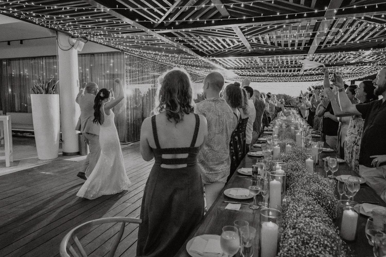 Black and white photo of wedding guests gathered around a long decorated table on an outdoor venue with string lights.