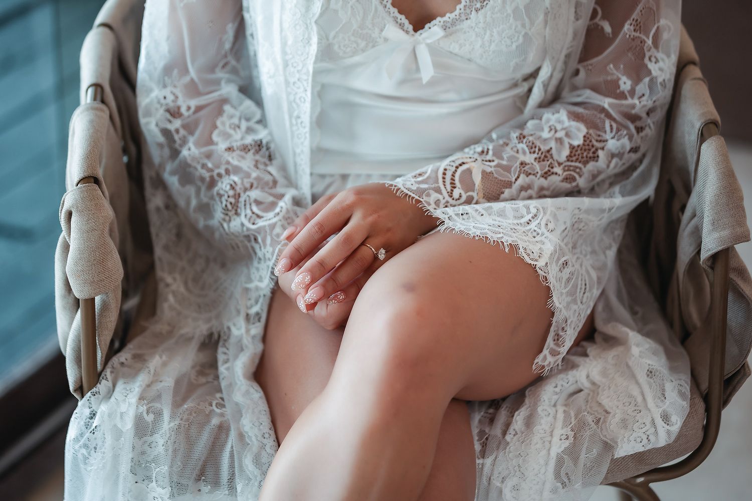 Bride in white lace robe sitting elegantly in a vintage chair, showcasing her engagement ring.