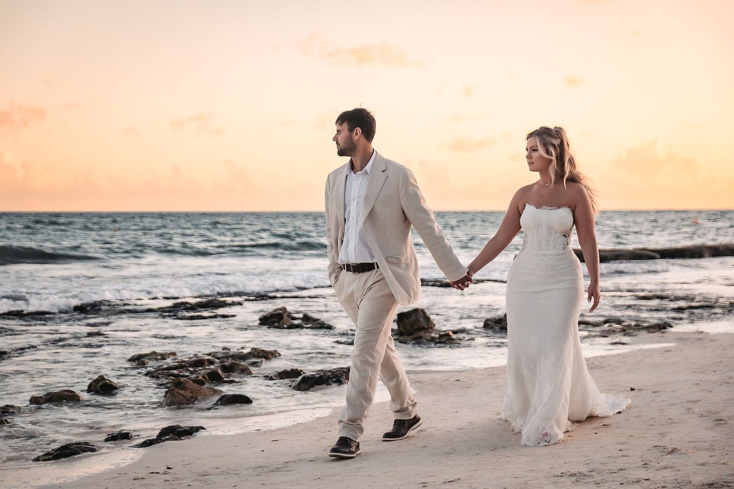 Bride and groom walk hand in hand on a sandy beach at sunset, wearing elegant white wedding attire.