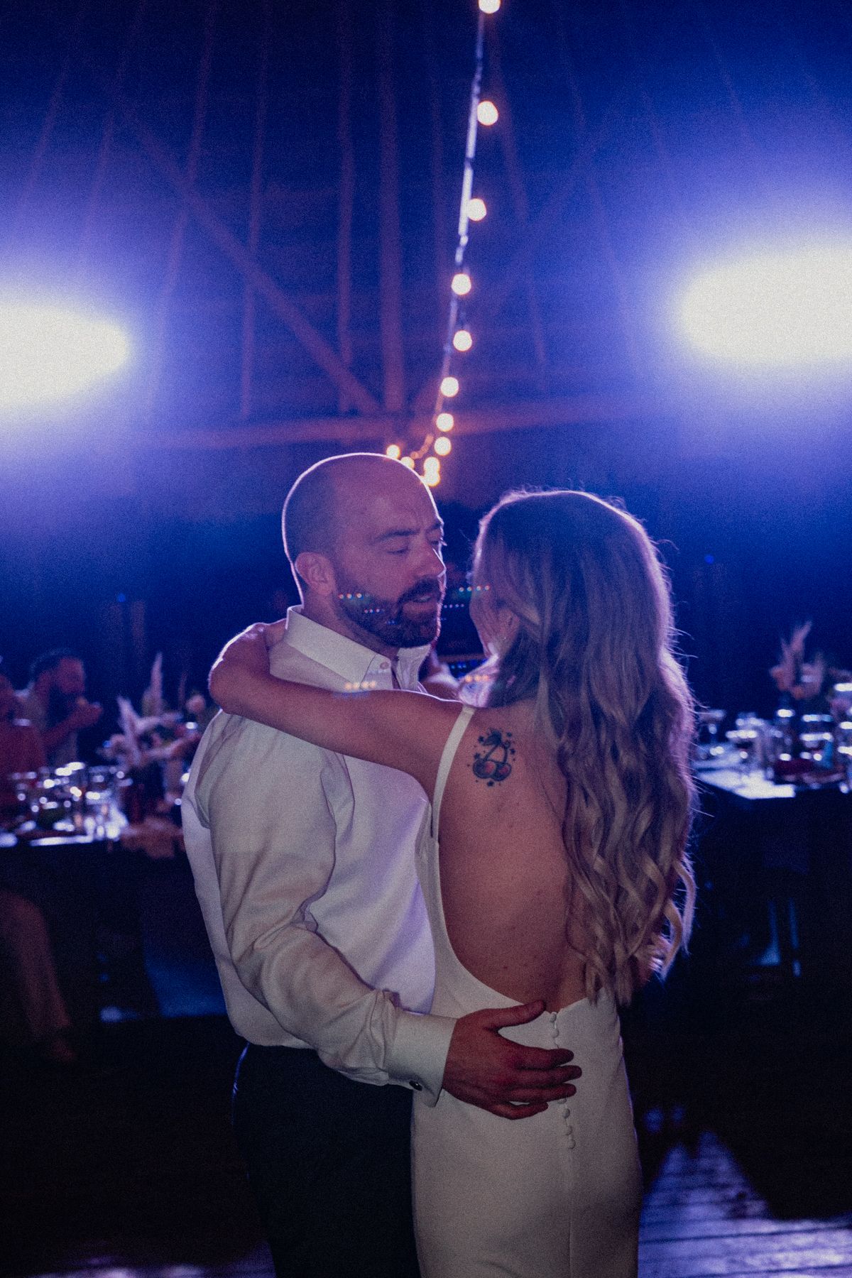 Couple shares intimate first dance under string lights with blue stage lighting at evening wedding reception.