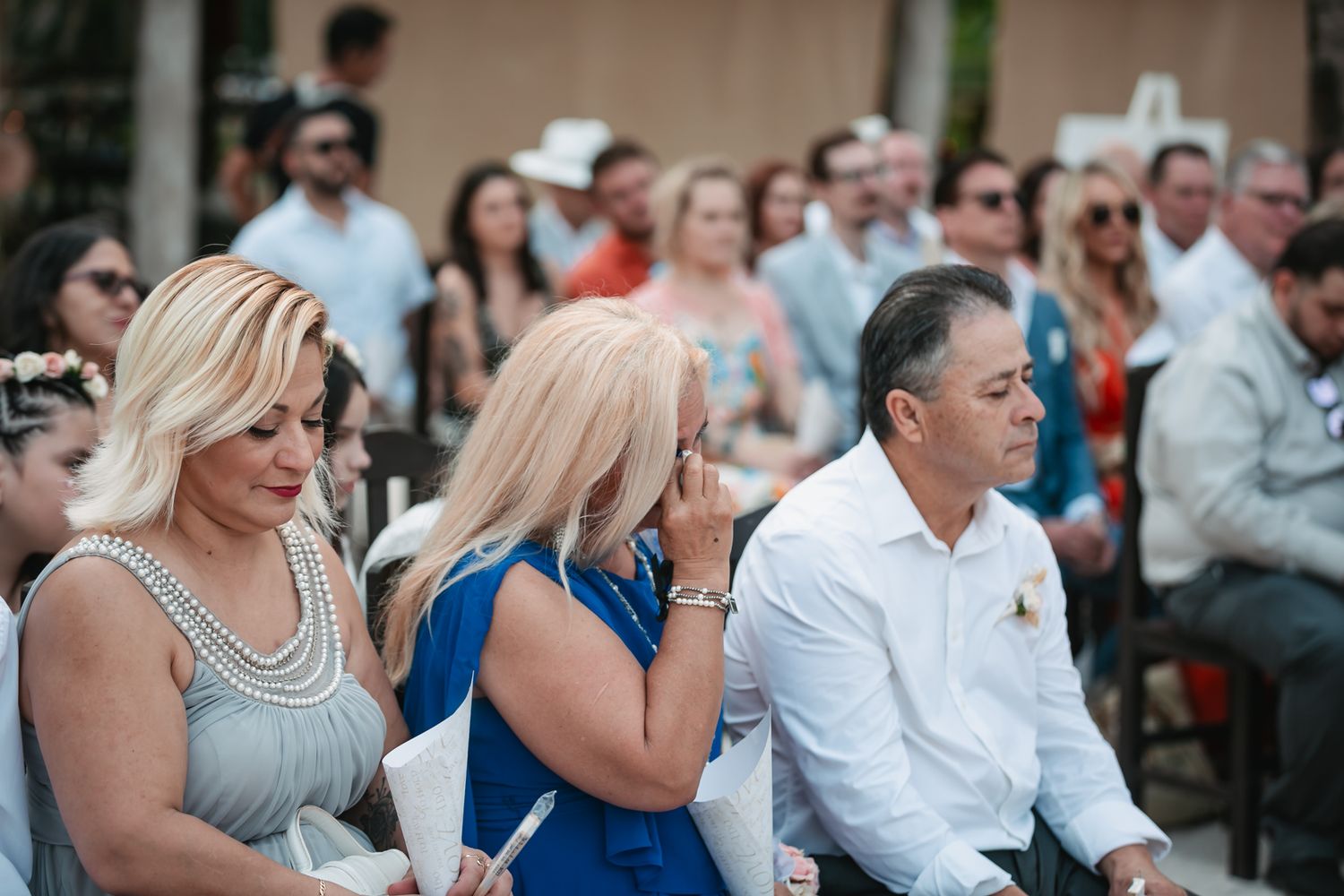 Emotional wedding guests seated at an outdoor ceremony, with a woman in blue wiping tears during the celebration.
