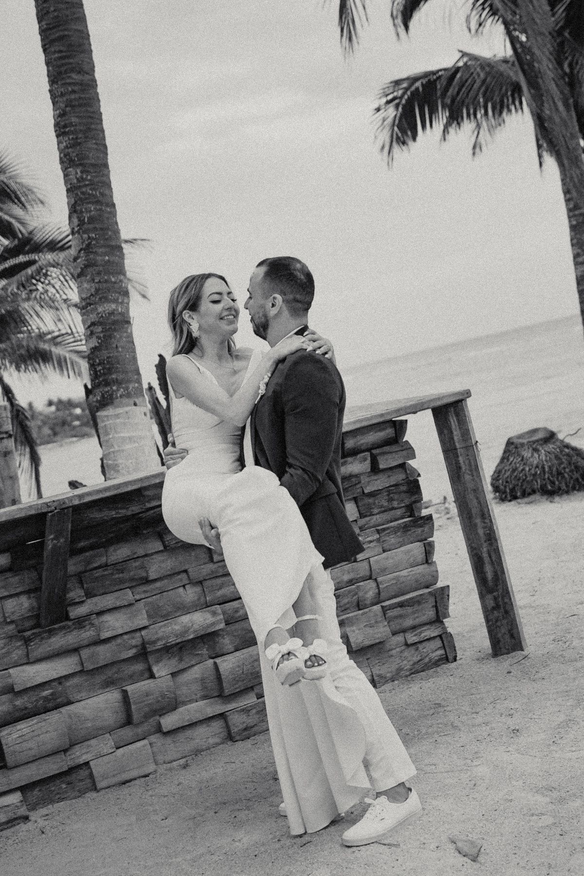 Black and white photo of bride seated on stone wall embracing groom near tropical beach.