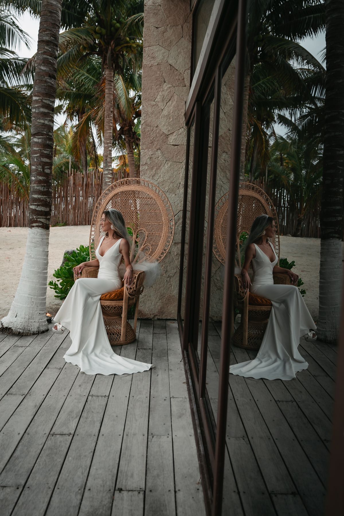 Bride in white gown sits in a wicker peacock chair on a wooden deck, reflected in glass, surrounded by palm trees.